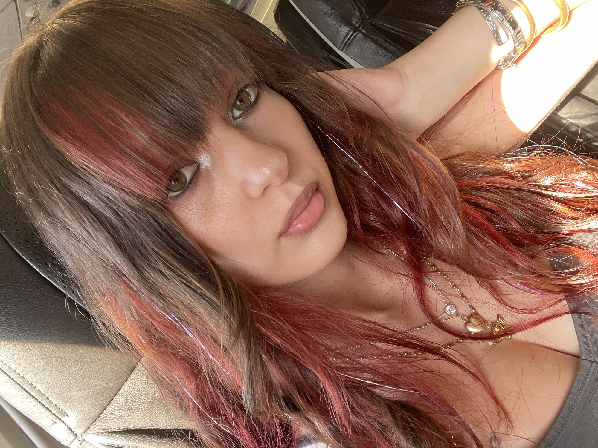 A medium close-up, high-angle selfie shows a young woman with long, wavy, dark brown hair with red highlights and tinsel strands. She has bangs and her hair falls around her shoulders. She is looking at the camera with her head tilted slightly and her right hand resting on her cheek. She is wearing a gray top and several necklaces, including a gold chain with a heart pendant and an angel charm, and a silver chain with a clear stone. She has bracelets on her left wrist. The background is blurry and appears to be the interior of a car, with a leather seat visible on the left and a darker seat on the right. Sunlight is casting shadows across the scene. The woman has full lips with glossy pink lipstick, and dark eyeliner. The overall lighting is warm and natural. There is no discernible location in the city of Unknown, Israel.