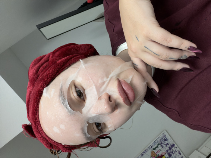 The scene depicts an individual in a close-up, upward-angled shot within an indoor setting in Ramat Gan, Israel. The subject's face is largely obscured by a white sheet mask with circular indentations, revealing open eyes that gaze directly forward and full lips colored with a muted rosy pigment. The subject's hair is contained by a dark red, textured head towel, from which damp, dark hair strands emerge at the top. The left hand of the subject is positioned against the jawline. This hand features long, pointed fingernails painted a dark maroon color, a ring on the left ring finger, and multiple dark linear tattoos on its dorsal surface. The subject wears a dark maroon long-sleeved garment with a white collar visible underneath. The background consists of a light-colored wall. A framed artwork, displaying colorful abstract or cartoonish elements, is partially visible in the upper left, while a dark-framed rectangular object, possibly a mirror, is partially visible in the lower right.
