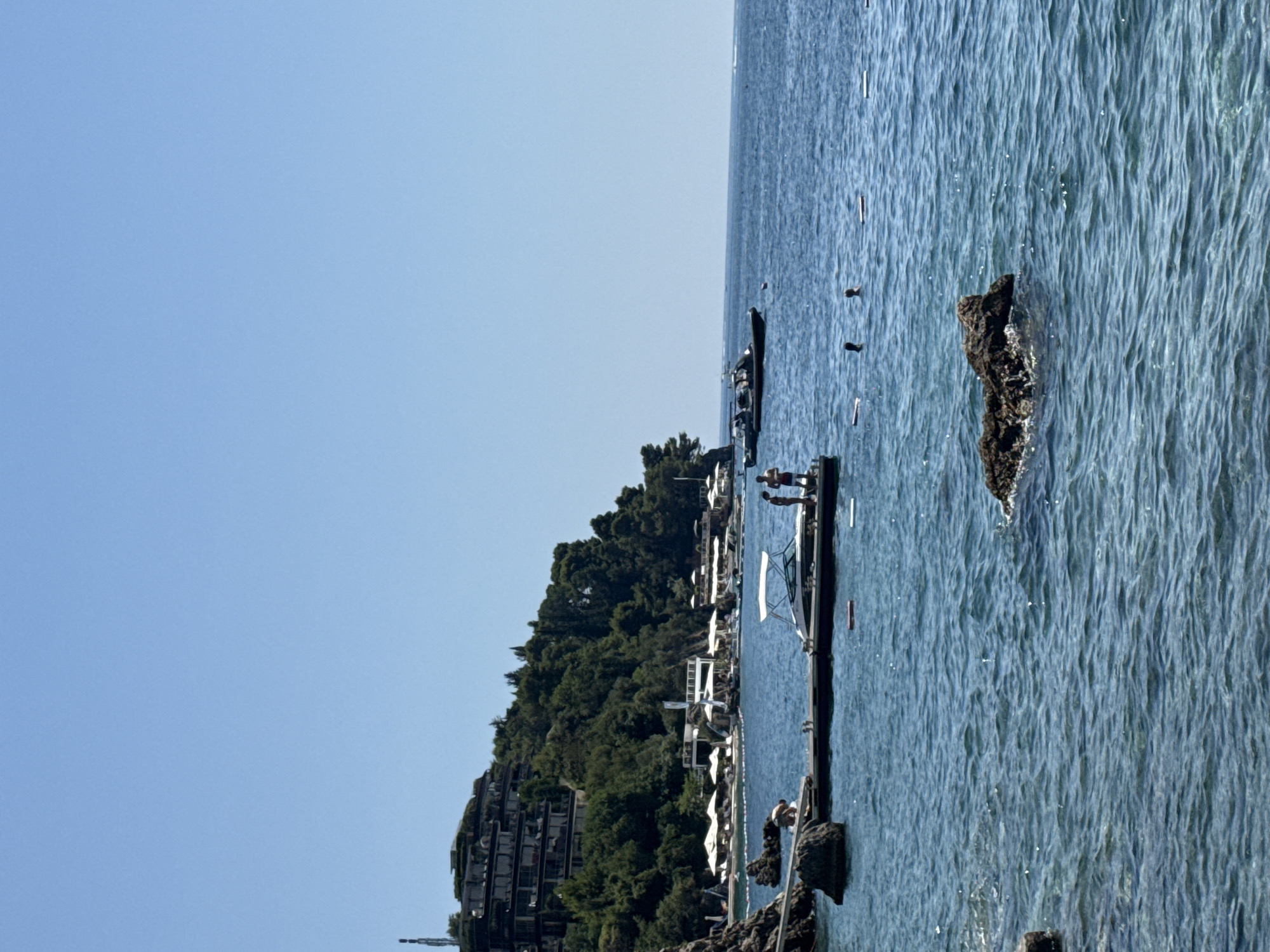 This image depicts a scenic view of the Adriatic Sea near Budva, Montenegro. The foreground shows clear blue water with gentle waves, and several rocks are visible, some partially submerged. A dark-colored rock formation is prominent in the lower right portion of the frame.
A wooden pier extends from the left side of the image into the water, with a white motorboat docked alongside it. Three people are standing on the pier near the boat, two appearing to be adults and one possibly a child. Another person is seated on a rock to the left of the pier.
In the mid-ground, a darker, larger boat with several individuals on board is sailing on the water. The coastline to the left features a lush, green hillside densely covered in trees. Several buildings, likely hotels or apartments, are visible cascading down the slope. Below the buildings, closer to the water's edge, are structures that appear to be part of a beach resort, with white umbrellas and lounge chairs arranged along the shore.
The background is dominated by a vast expanse of clear, light blue sky, with no clouds visible. The overall scene suggests a bright, sunny day at a coastal resort.