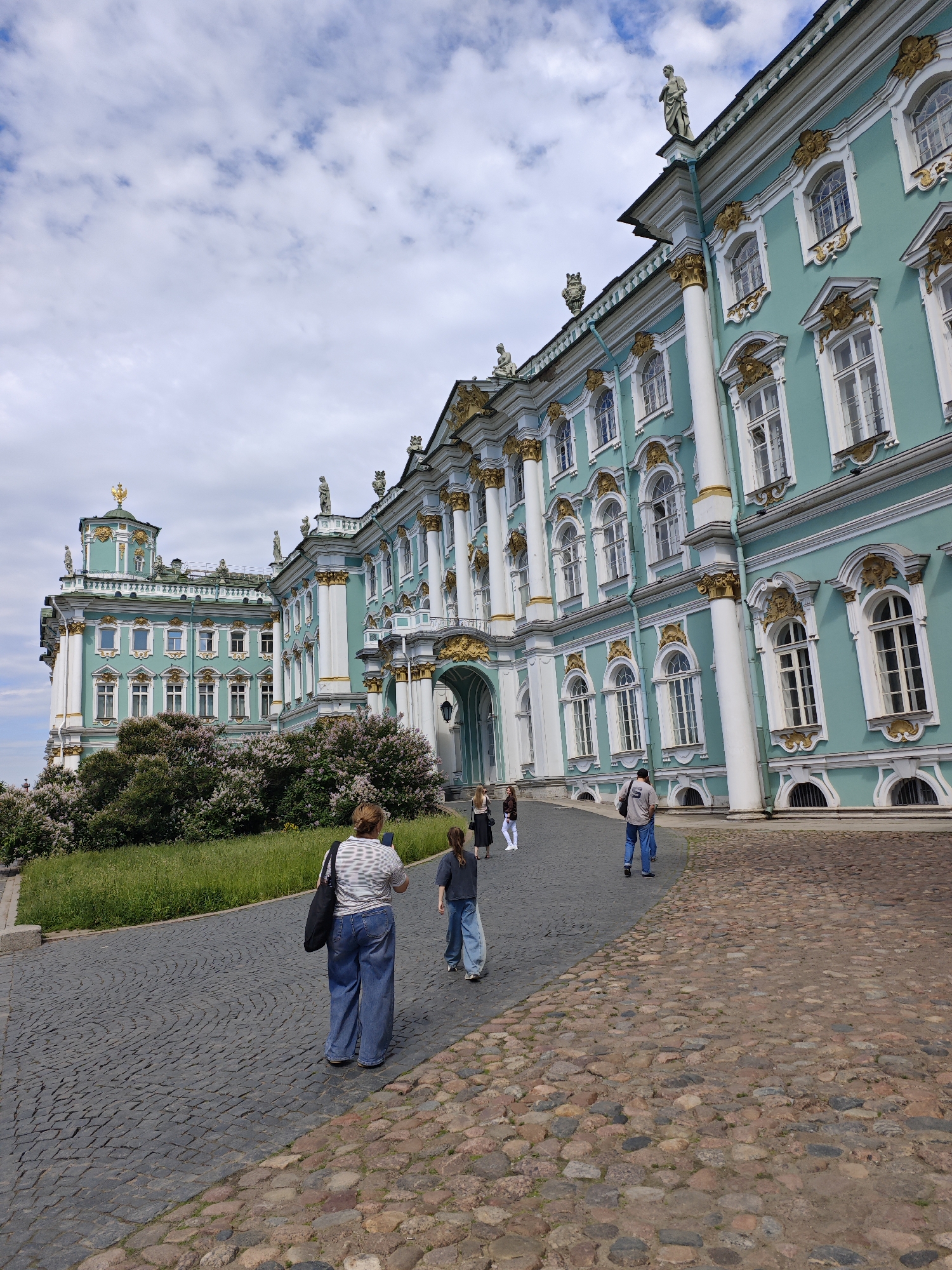 The image shows a wide cobblestone path leading to a grand, teal-colored building with white columns and gold accents. The building appears to be a historic palace or museum, with ornate windows and statues adorning its facade. A row of blooming trees with white flowers lines the left side of the path. Several people are walking along the path, some facing away from the camera and towards the building, others walking in the opposite direction. The sky is partly cloudy, with a mix of white clouds and blue sky. The overall impression is one of a bright, sunny day in a picturesque historical setting, likely in Saint Petersburg, Russia, given the architectural style.