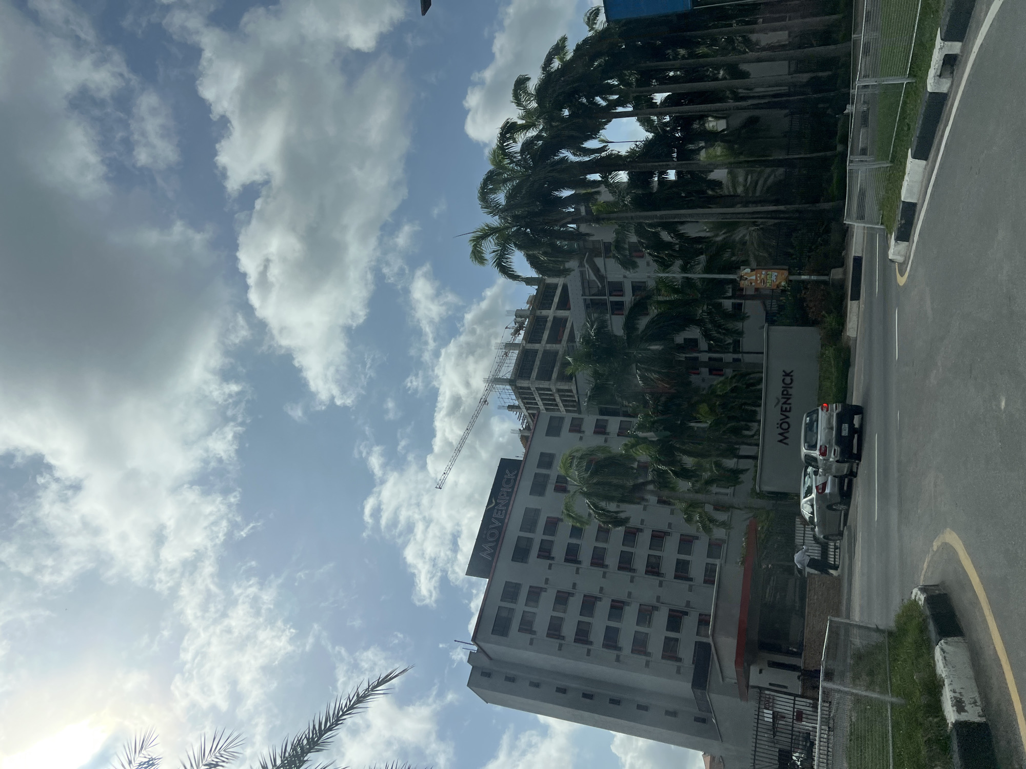 The image shows a street scene in Ikoyi, Nigeria, on a bright day with a partly cloudy sky. The Movenpick Hotel is visible on the left, along with another building under construction. Several cars are parked on the side of the road, and a man is standing near one of the vehicles. Palm trees line the road, and a sidewalk is present.