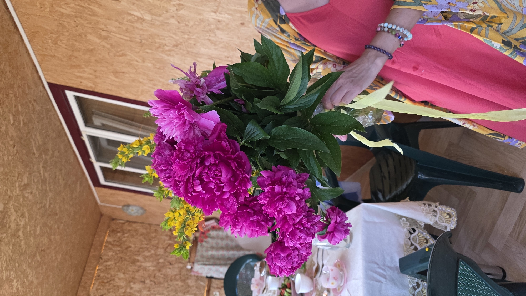 This media file shows a close-up of a bouquet of bright pink peonies and yellow flowers. The bouquet is held by a person wearing a vibrant pink dress and a yellow patterned kimono-style robe. The person's right arm is visible, adorned with beaded bracelets. The background suggests an outdoor or semi-outdoor setting, possibly a porch or covered patio, with wooden paneling on the walls and ceiling. A window with white trim is visible in the background.
To the right of the bouquet, a white tablecloth with ornate gold trim is draped over a table, on which white teacups and saucers are arranged, hinting at a tea party or a celebratory gathering. A dark green plastic chair is partially visible, suggesting a casual outdoor setting. The lighting appears to be natural daylight, likely mid-day. The overall mood conveyed is cheerful and summery, with the bright colors of the flowers and clothing contributing to this feeling.
The location is identified as Kupoyarovo, Russia. There is no visible text in the image. The activity taking place is the presentation or arrangement of a flower bouquet, possibly in preparation for an event.