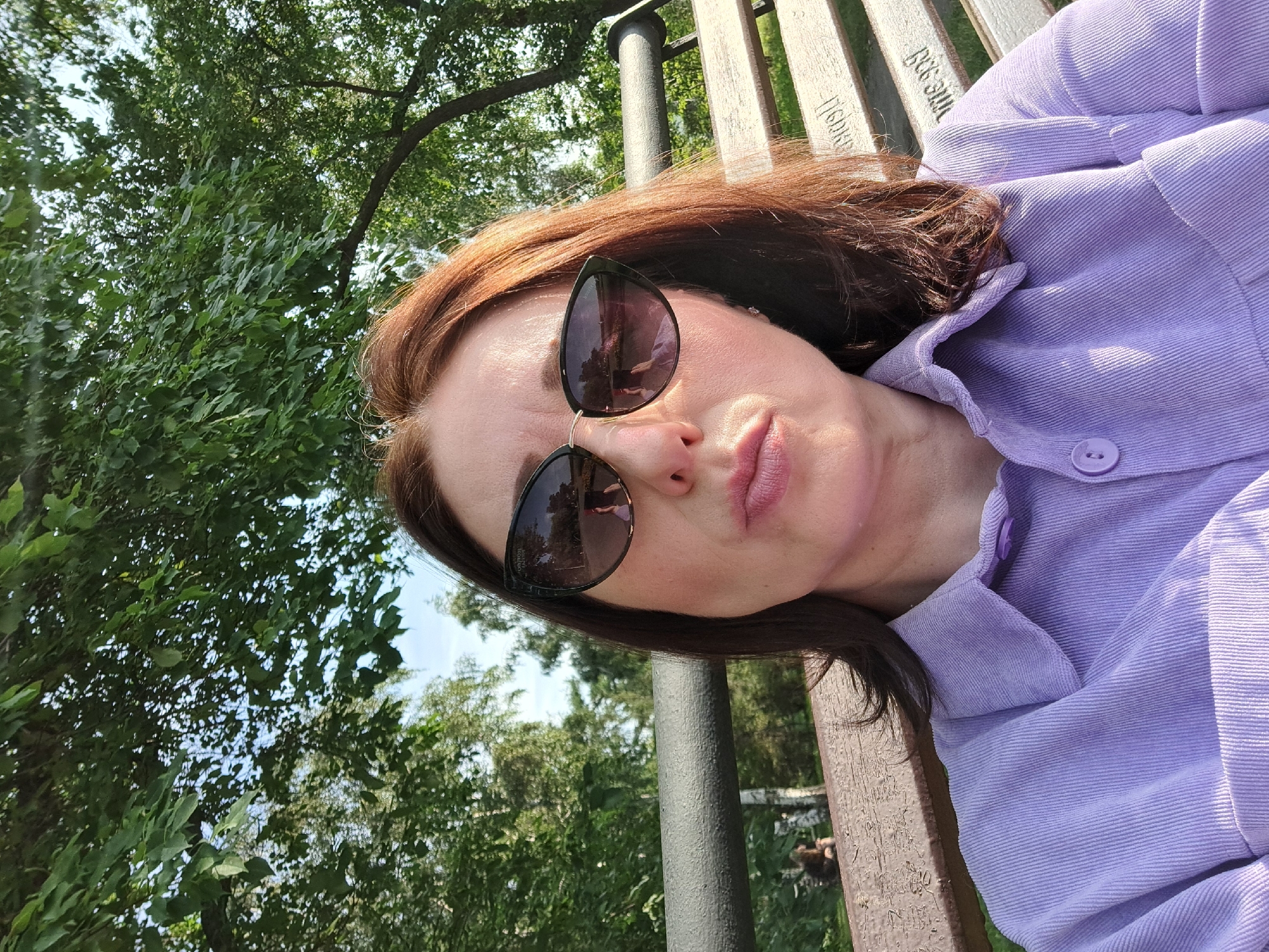 A woman with auburn hair, wearing sunglasses and a lavender corduroy shirt, is seated on a park bench. She is looking towards the camera with her lips pursed. The bench is wooden with metal armrests and appears to be inscribed with text. In the background, there are trees and foliage, suggesting an outdoor park setting. The lighting indicates it is daytime, possibly late afternoon due to the warm tone of the light. The scene is devoid of any specific landmarks that would pinpoint its location within the city of Irkutsk, Russia.