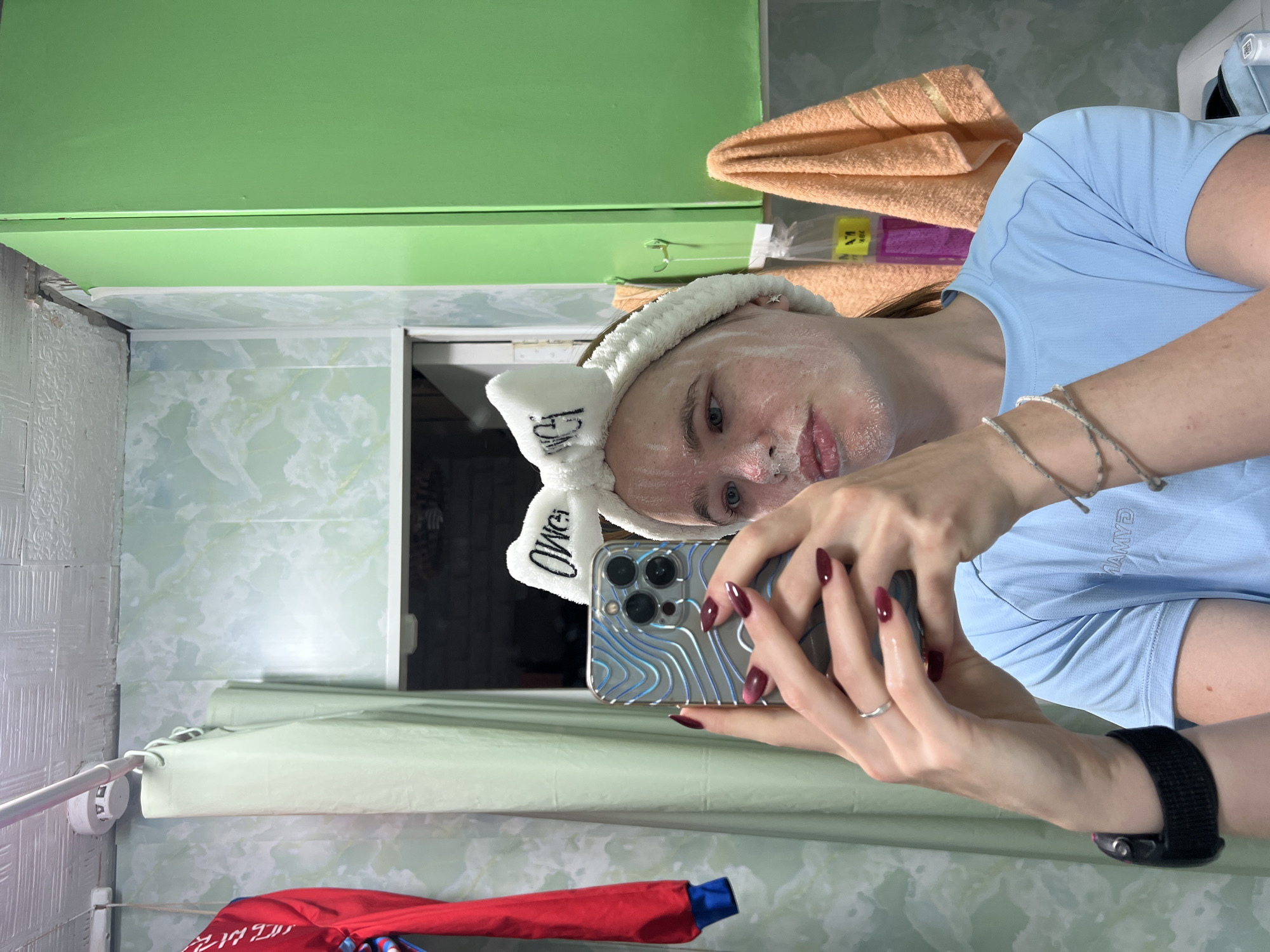 A young woman is taking a selfie in a bathroom. She is wearing a white headband with the word "OWEI" embroidered on it in black, and has a white cream mask on her face. She is holding a smartphone in her hands, with her reflection visible on the screen. The bathroom walls are covered in a green marble-patterned wallpaper, and there are green cabinets on the right side of the image. An orange towel is hanging on a hook to the right of the cabinets. A red jacket with blue trim is hanging on the left side of the image. The ceiling appears to be made of white tiles, and a smoke detector is visible on the left. The overall impression is of a small, functional bathroom, likely in a residential setting. This scene is located in the city of Chaikovsky, Russia.