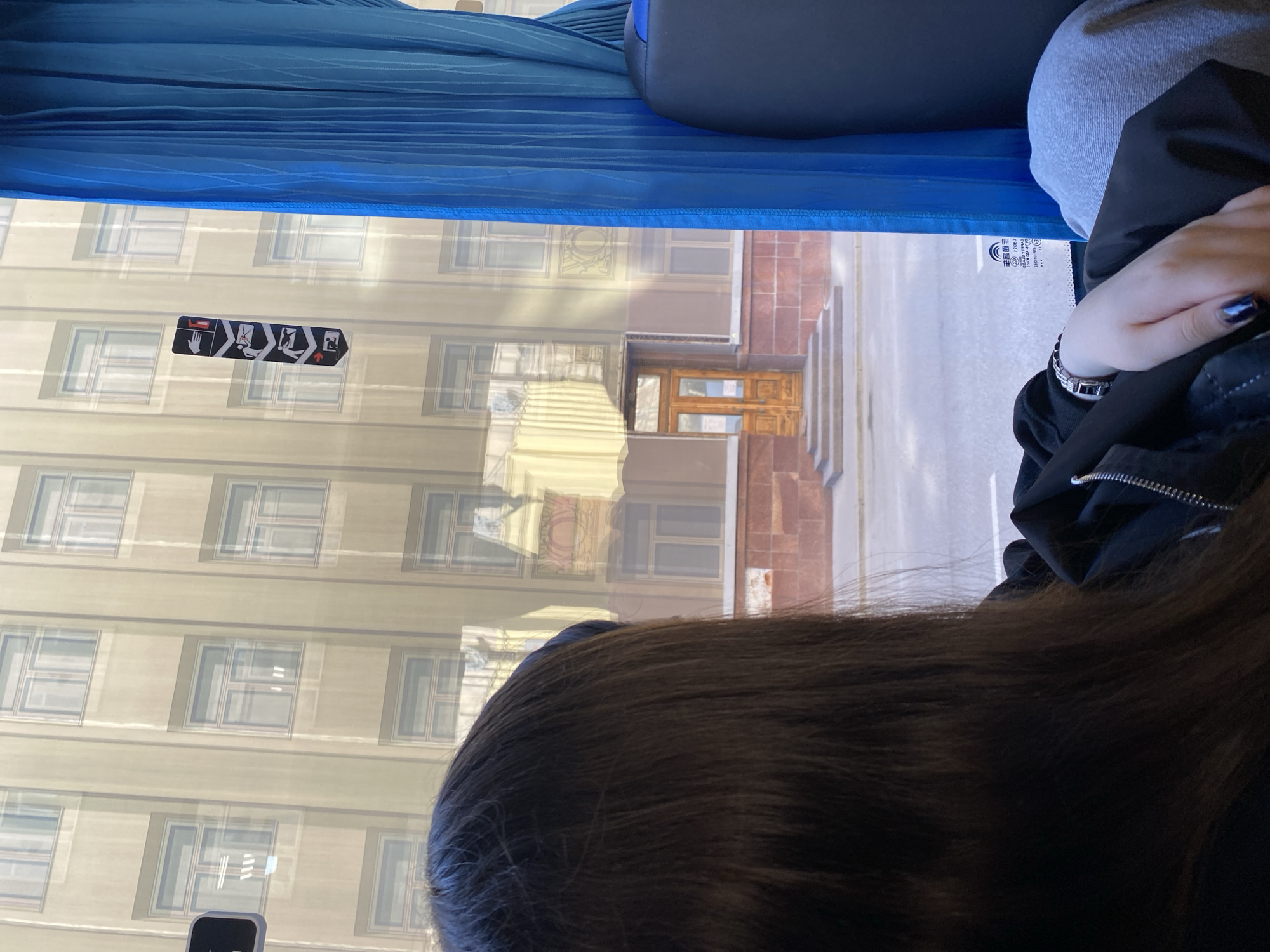Here is a detailed description of the image:
**Scene:** The image appears to be taken from inside a bus or other vehicle, looking out of the window. A person's head and shoulder are visible in the foreground. The view through the window shows a street scene with buildings.
**People:**
* A person with dark hair is partially visible in the foreground. Their hand, adorned with a bracelet and blue fingernails, rests on what appears to be a jacket.
**Objects:**
* Bus window.
* Blue curtain.
* Seat upholstery (blue and gray).
* Building with multiple windows, likely an administrative or residential building.
* Building entrance with double doors and steps.
* Bus safety sticker.
* Bracelet.
* Jacket.
**Actions:**
* The bus/vehicle is likely in motion, as the scene outside is visible through the window.
* The person is looking out the window.
**Environment:**
* Daytime.
* Urban environment.
* The architectural style of the building suggests a location in Moscow, Russia.