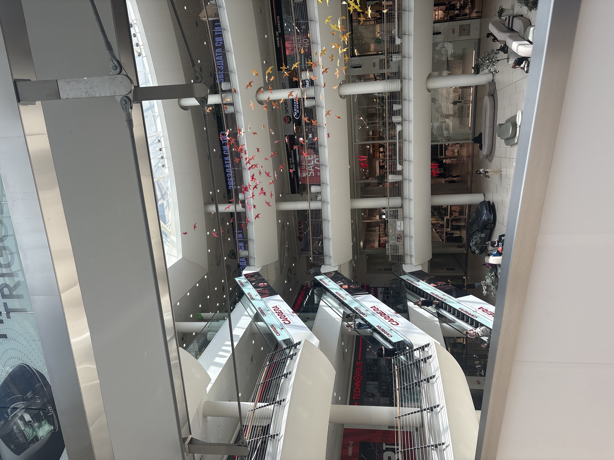 This image shows the interior of a multi-story shopping mall, possibly in the city of Unknown, Unknown. The view is looking down from a high vantage point, capturing several floors of shops and escalators. A decorative installation of red and orange bird-shaped decorations is suspended in the open space between the floors. Escalators are visible, marked with "CARRERA" branding, and people are riding them. The shops have displays and advertisements, including phrases like "MID SEASON SALE" and brands like "H&M". Seating areas with modern furniture and potted plants are placed near shops, with people sitting and standing around. The architecture of the building features white columns, beams, and glass panels, providing natural light. The overall scene depicts a typical commercial environment within a modern shopping center.