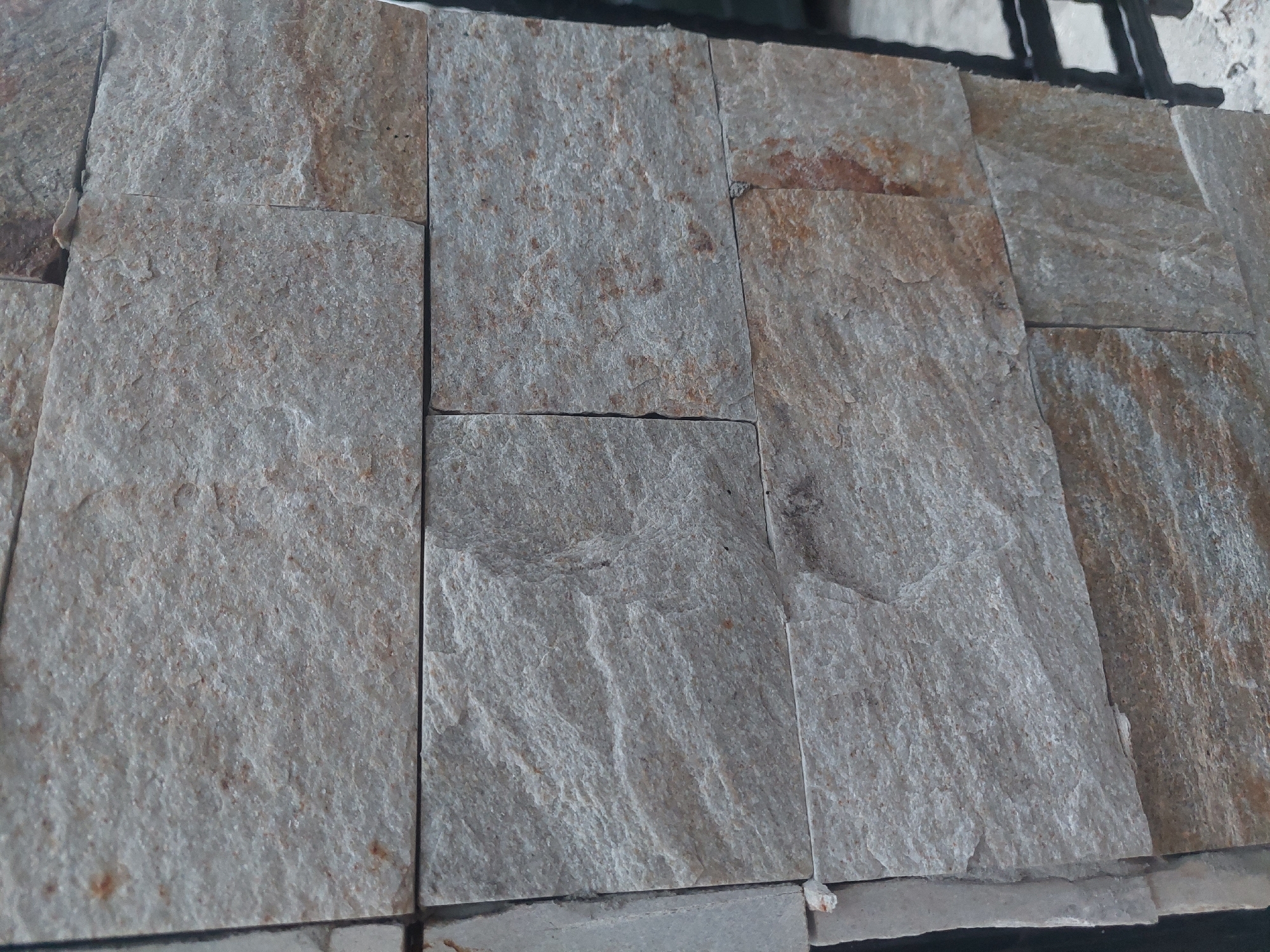 A textured stone wall, likely a facade, is shown in a close-up, vertical shot. The stones are rectangular, arranged in horizontal rows, with varying shades of grey and brown, and a rough, natural texture. Some stones have visible striations and flecks of darker minerals. The mortar between the stones is a lighter grey. In the background, a dark, possibly black, metal structure is visible on the right side of the image, suggesting it could be a fence or gate. The overall impression is of an outdoor setting, possibly a building exterior or garden feature. The location is not specified in the image itself, and therefore the mention of Kostenets, Bulgaria, is extraneous to the visual information provided by the image.