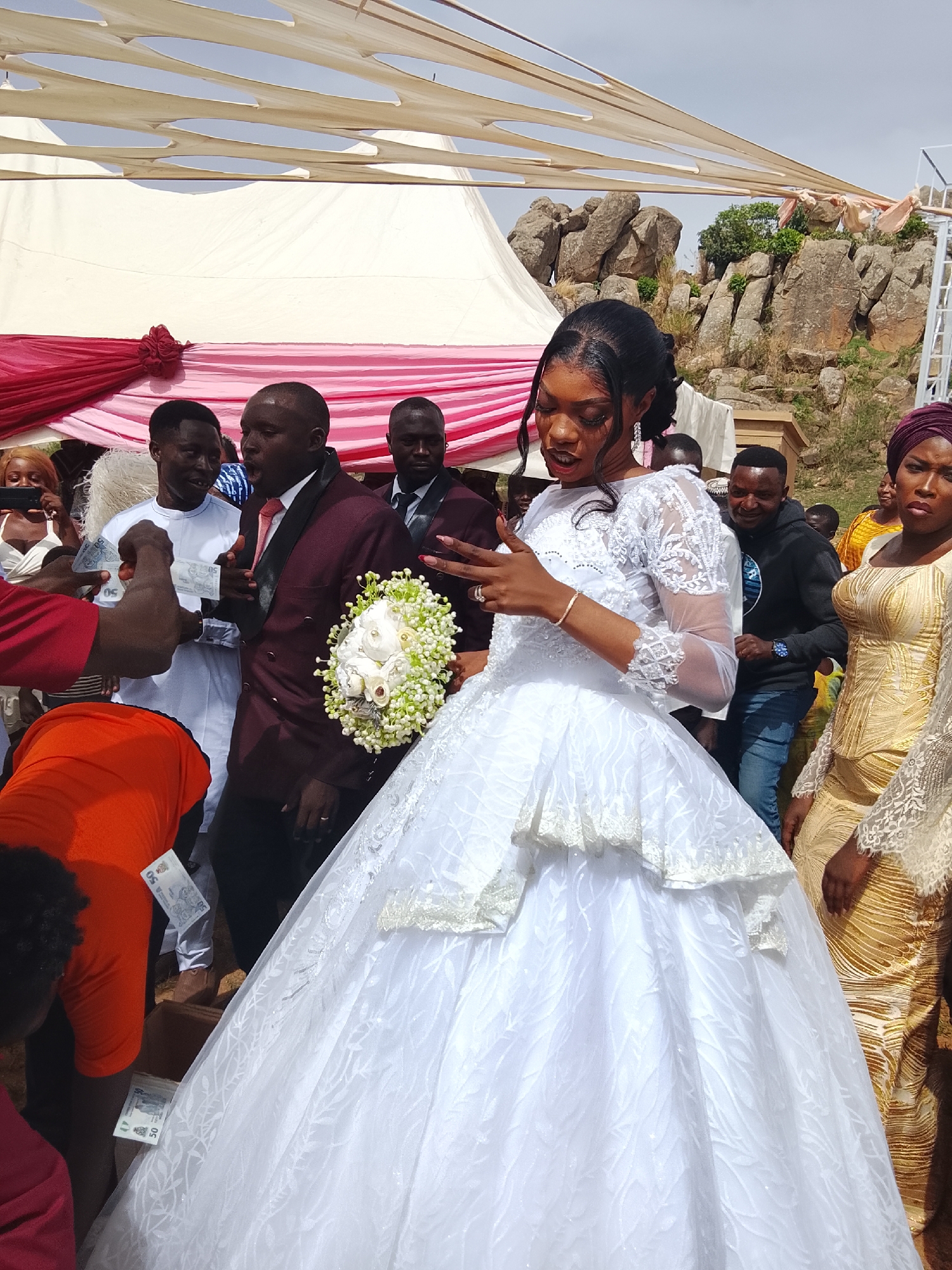 Dogon Dinia, Nigeria: A wedding ceremony is taking place outdoors. A woman in a white wedding dress holds a bouquet of white flowers. Men in suits and a woman in a gold dress stand nearby. One man is handing money towards the bride. Others in the background are observing. A tent structure is overhead. Behind the tent are rock formations.