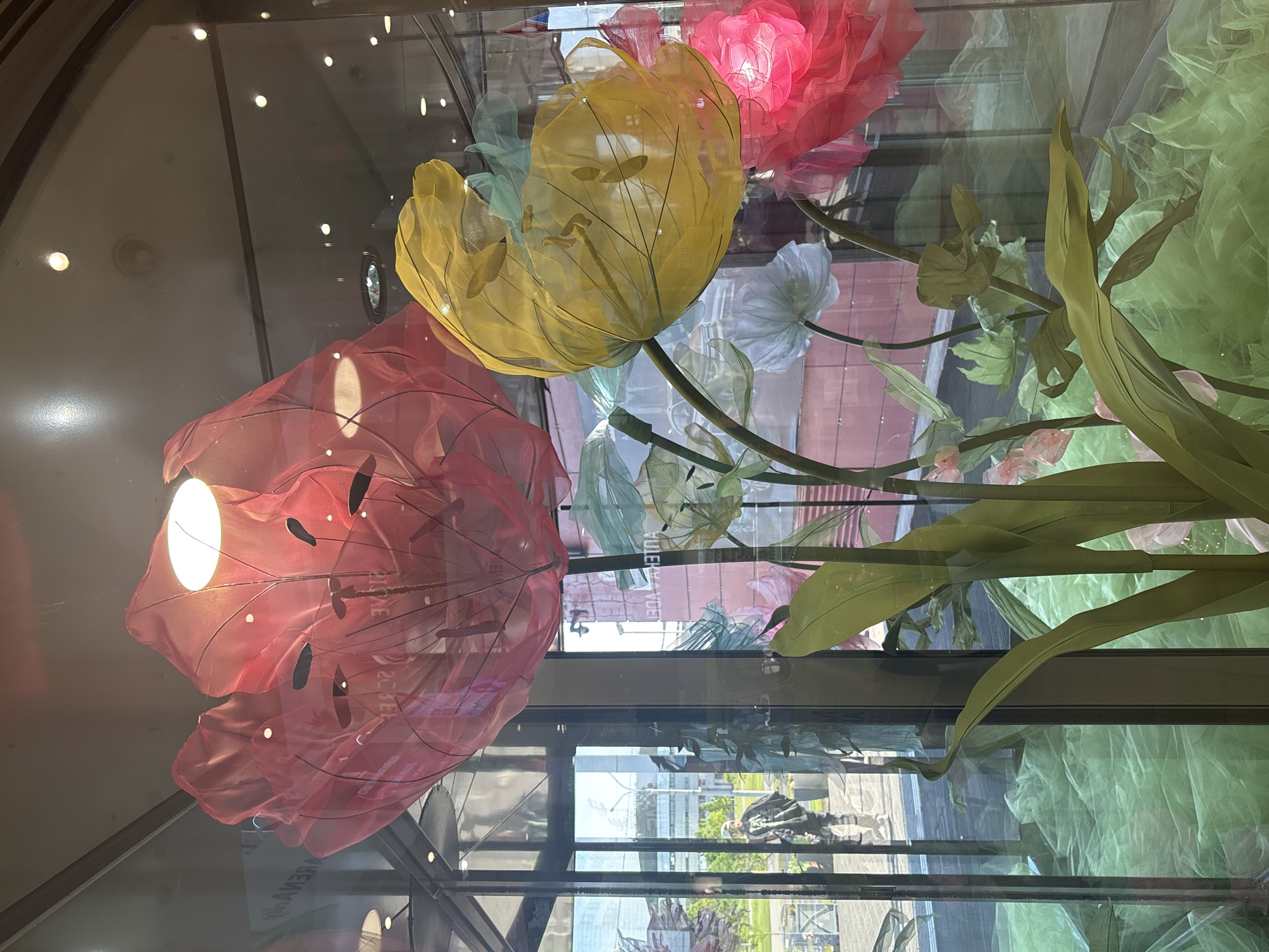 The image displays a vibrant and luminous arrangement of large, artificial flowers, viewed through a clear glass enclosure. The exhibit features several oversized blossoms crafted from translucent, sheer fabric, possibly organza or silk, supported by visible wire frames. A prominent red flower, illuminated from within by a bright light source, dominates the left side of the composition. A yellow flower is positioned centrally, and a pink one appears towards the right, also glowing. Green artificial leaves and stems are interspersed throughout the floral display.
The setting appears to be an indoor public space, such as a lobby or a large display window, in Minsk, Belarus. The glass surface intensely reflects the external environment, showcasing modern building facades, a bright sky, and the silhouette of a person walking by in the lower right portion of the reflection. These reflections indicate it is daytime.
The display creates an ethereal and decorative ambiance due to the internal illumination of the colorful flowers. Several pieces of reversed text are visible in the reflections on the glass. One distinct reflection reads "REVIVER," and another appears to be "VUIEAYDEI," likely elements of nearby signage or advertisements.