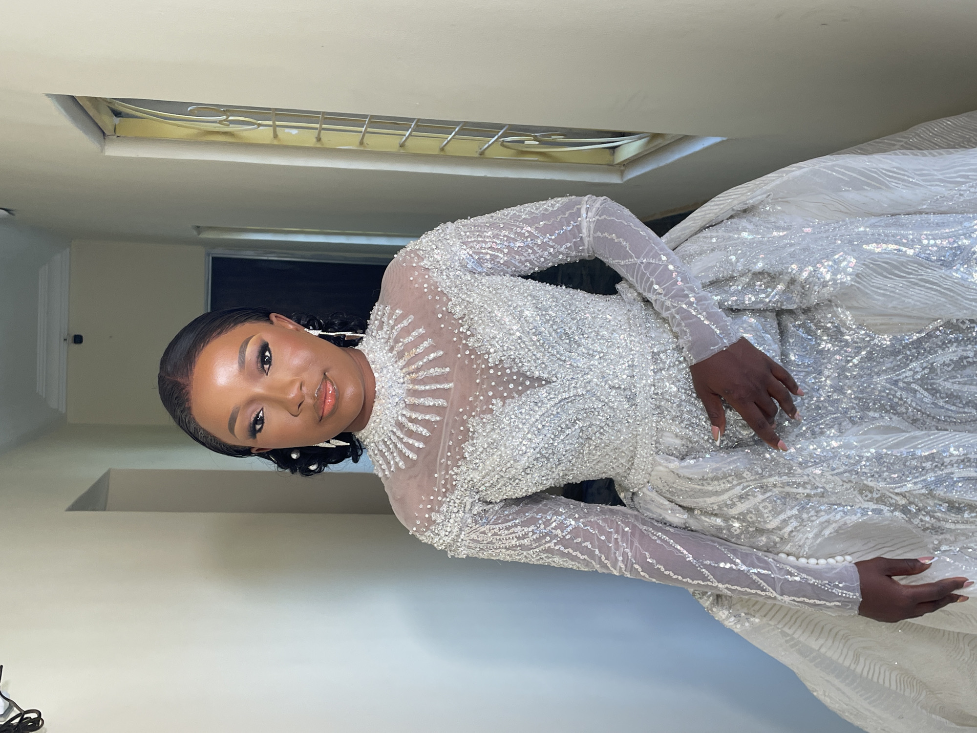 A woman with dark skin and neatly styled black hair is wearing a heavily embellished white and silver long-sleeved dress, which appears to be a wedding dress. She stands inside a room with cream or light beige walls. There is a rectangular recessed light fixture with a metal grill in the ceiling above her. The woman's face is in focus and she is looking at the camera. The location of this image is not evident.
