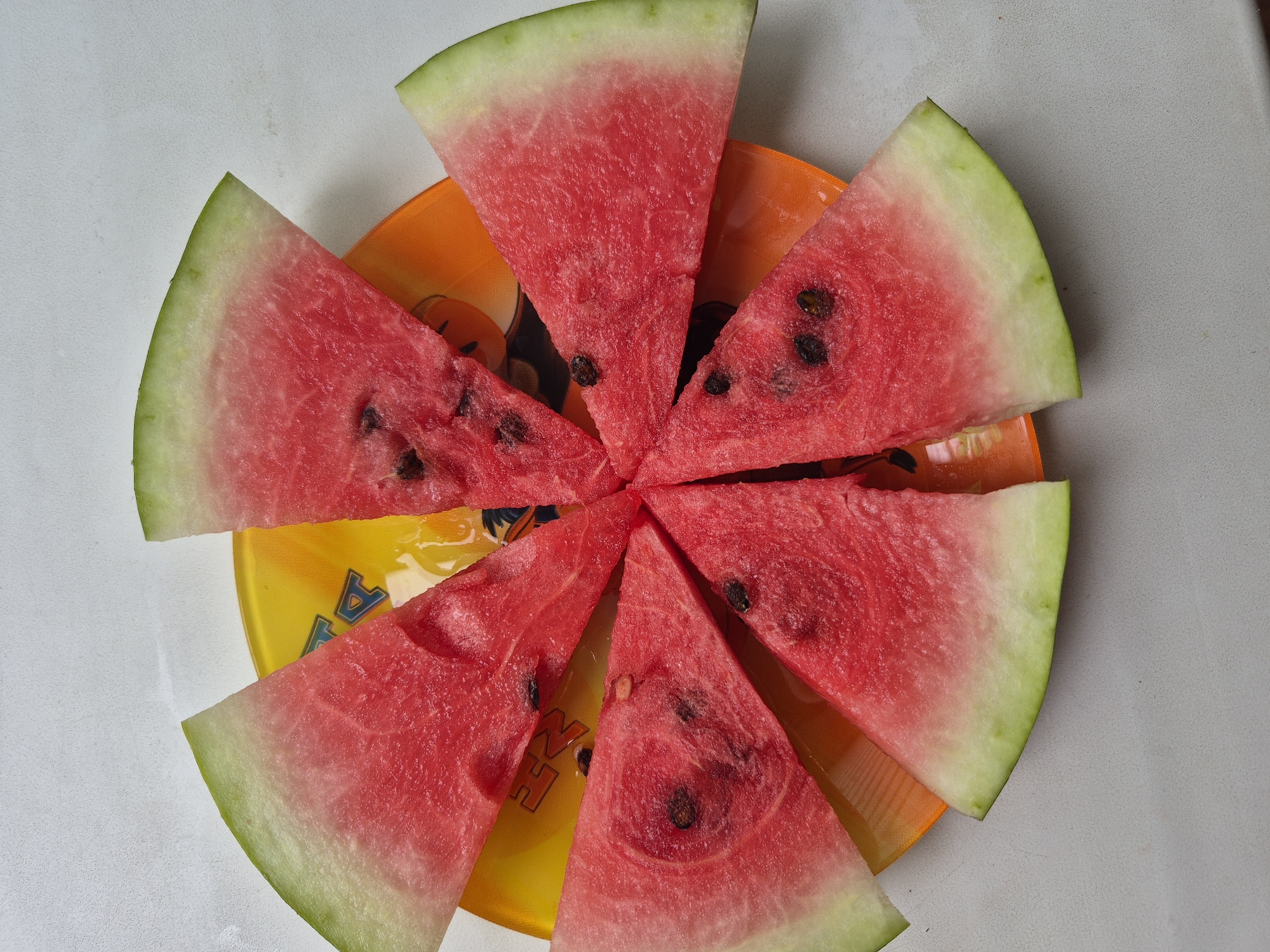 Eight triangular slices of watermelon are arranged in a circular pattern on a yellow and orange patterned plate. The watermelon slices are vibrant red with visible black seeds and a light green rind. The plate itself has a subtle wave-like design and appears to be made of plastic or glass. The scene is captured from a top-down perspective against a plain white textured surface. There is no information available to associate this image with the city of Kokshetau, Kazakhstan.