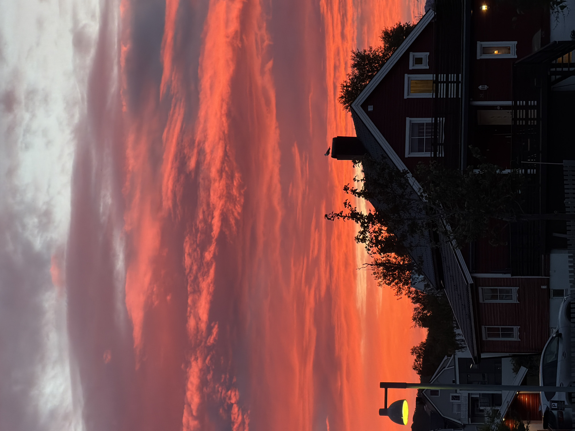This is a vertical, eye-level, full shot of a dramatic sunset in Harstad, Norway. The sky is a vibrant display of reds and oranges, with wispy clouds streaking across the scene, tinged with purple and gray. In the foreground, a dark red wooden house with white trim is visible. The house has multiple windows, some illuminated, and a dark-colored porch with railings. A seagull is perched on top of a chimney on the roof. To the left of the house, a street lamp with a yellow light casts a warm glow. In front of the house, a silver SUV is parked. Other houses and trees are silhouetted against the colorful sky in the background.