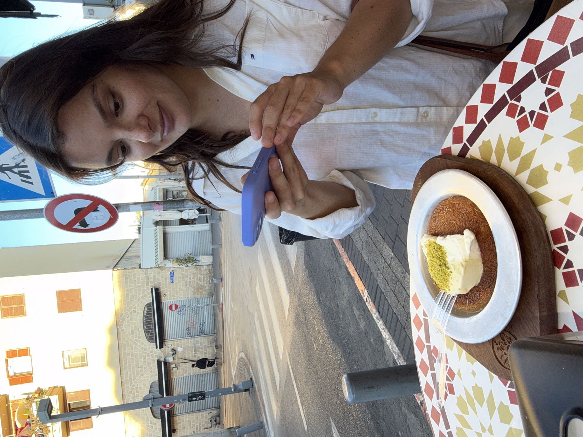A young woman with dark hair is taking a photo of a dessert with her phone in Tel Aviv, Israel. The dessert is a plate of Kunafa, a traditional Middle Eastern pastry, topped with a scoop of ice cream and pistachios. It is served on a wooden board on a table with a colorful geometric pattern. The woman is wearing a white linen shirt. In the background, a street scene is visible with buildings, a pedestrian crossing, and street signs. The sky is clear and blue.