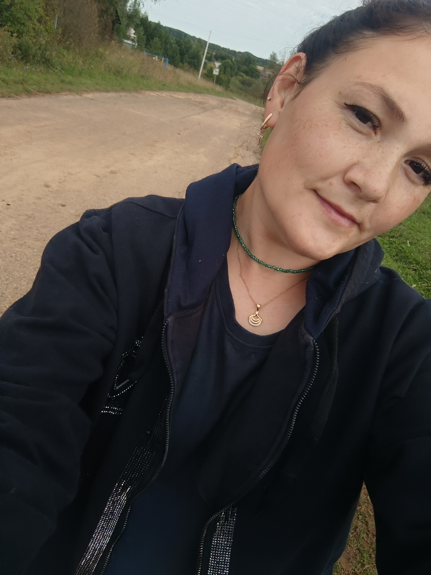 A young woman with dark hair pulled back and freckles on her face is pictured from the chest up. She is wearing a dark blue hooded jacket over a dark blue t-shirt, and a green beaded necklace with a gold pendant. She is also wearing gold earrings. The woman is looking towards the camera with a slight smile. She is outdoors in what appears to be a rural or suburban setting. A dirt road is visible behind her, with grassy areas and trees on either side. In the distance, there are rolling hills and a cloudy sky. This image was taken in Karsha, Russia.