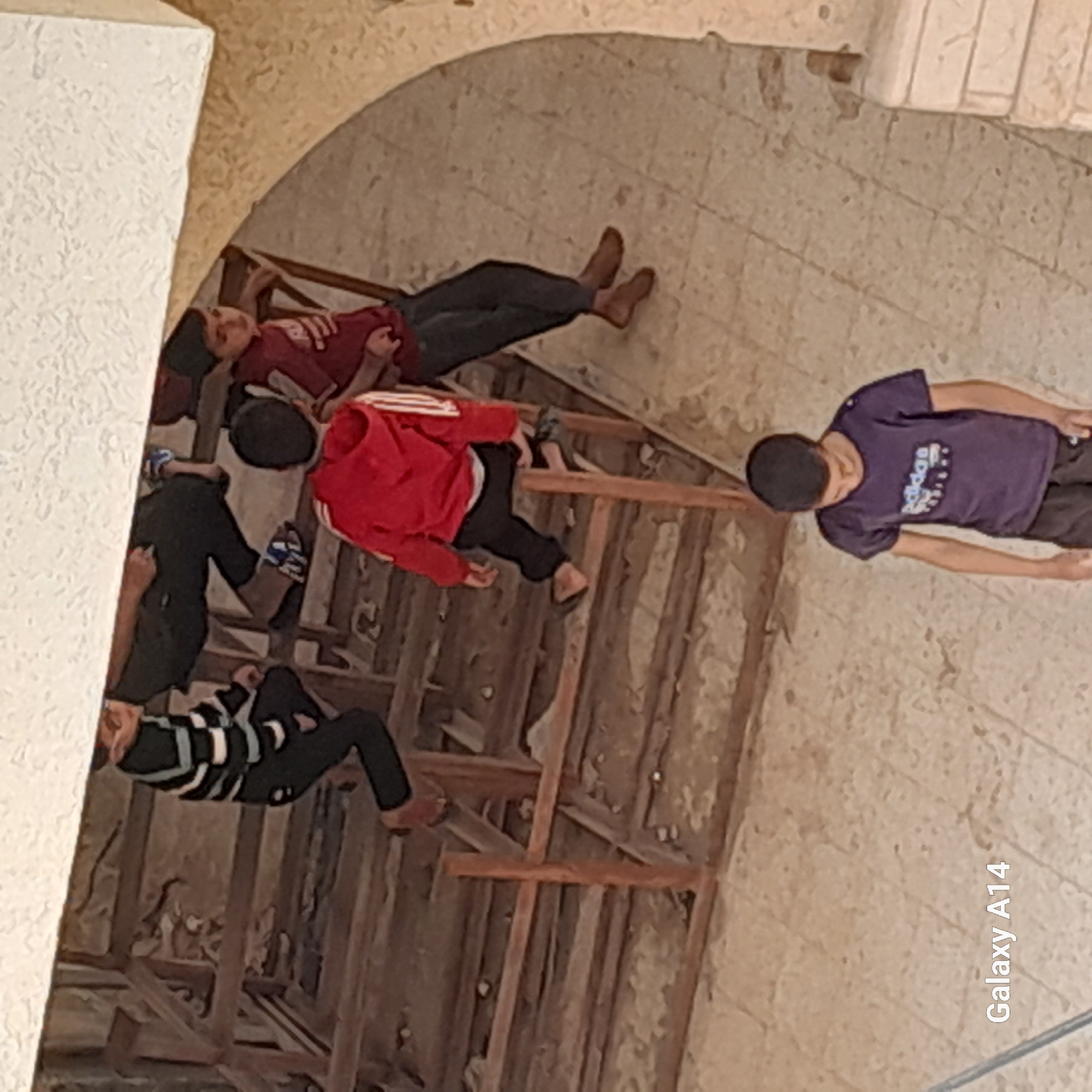 The image captures a scene featuring several children in what appears to be an indoor or semi-enclosed space. The setting includes a makeshift wooden structure, possibly scaffolding or a temporary platform, over a tiled or concrete floor. The walls also appear to be made of light-colored, textured material, possibly unfinished plaster or concrete.
There are at least five children visible in the image, most of whom are lying down or resting.
* One child, wearing a dark red long-sleeved shirt and dark pants, is lying on the upper part of the wooden structure, facing upwards.
* Another child, wearing a red hoodie and dark pants, is lying on a lower section of the wooden structure, facing sideways.
* A third child, wearing a purple t-shirt with a white design and dark pants, is lying on the tiled floor to the right of the wooden structure, facing upwards.
* A fourth child, wearing a black and white striped long-sleeved shirt and dark pants, is lying on the wooden structure on the left side of the image, facing sideways.
* Part of a fifth child, wearing a dark shirt and blue patterned arm sleeves, is visible near the top left of the wooden structure.
The children appear to be resting or relaxing. Their clothing is casual. The overall impression is one of a quiet, possibly afternoon, rest period in an unadorned or partially constructed environment. The lighting is soft and even, suggesting it is not direct sunlight.
A notable detail is the "Galaxy A14" watermark visible in white text in the bottom right corner of the original image, indicating the device used to capture the photo.