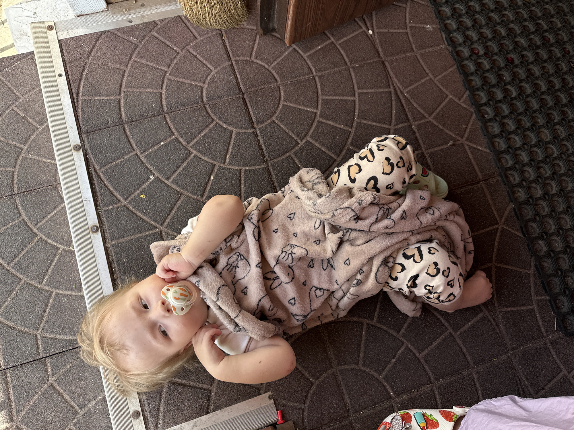 A toddler lies on a circular-patterned stone surface, possibly a patio or walkway, adjacent to a metallic threshold and a dark-colored wall or structure. The toddler is fair-skinned with blonde hair and is wearing animal-print pants and a light-colored blanket patterned with bunnies and hearts. They have a pacifier in their mouth and are looking towards the camera. Part of a dark mat is seen at the edge of the frame. The location is possibly in Nizhny Novgorod, Russia, though there is no definitive visual confirmation.