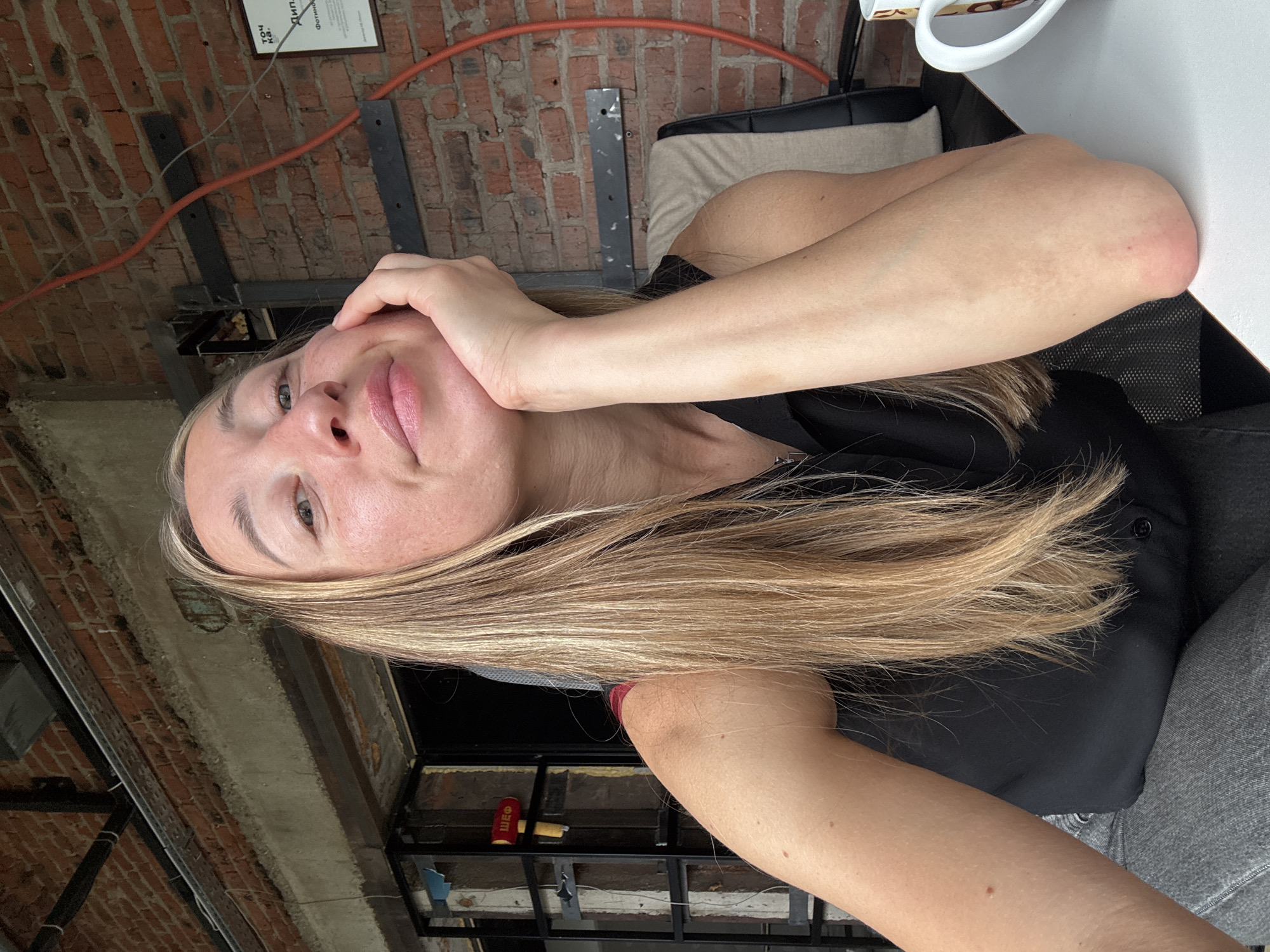 The media file is a close-up, portrait-oriented photo of a young woman. She is looking directly at the camera with a neutral expression. Her blonde hair cascades down her shoulders. Her left hand is gently touching her face, near her cheek and temple. She is wearing a black top and gray pants. The background suggests an indoor, possibly industrial or loft-style setting, with exposed brick walls and visible metal structures. There is a red electrical conduit running along the brick wall. Part of a certificate or document is visible on the wall in the upper left corner, with some Russian text on it. The lighting appears to be natural, coming from a window or overhead source, suggesting daytime. The overall mood is calm and informal. There is no indication of specific weather conditions or a particular event taking place; it appears to be a candid or posed portrait.