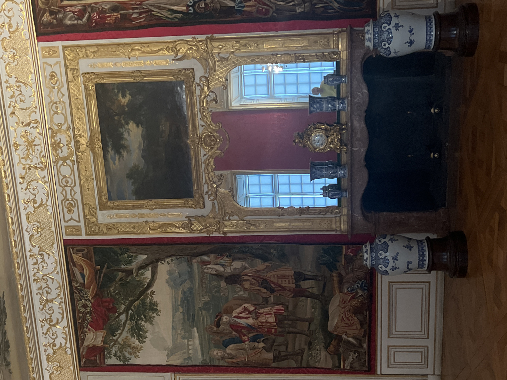 This image shows a room in the New Schleissheim Palace, located in Oberschleissheim, Germany. The room is ornately decorated with gold trim and features large tapestries and a painting above a fireplace. A person is visible in the reflection of a mirror above the fireplace, wearing a yellow shirt. Two large ceramic vases with blue and white patterns stand on wooden pedestals on either side of the fireplace. The floor is made of polished wood.