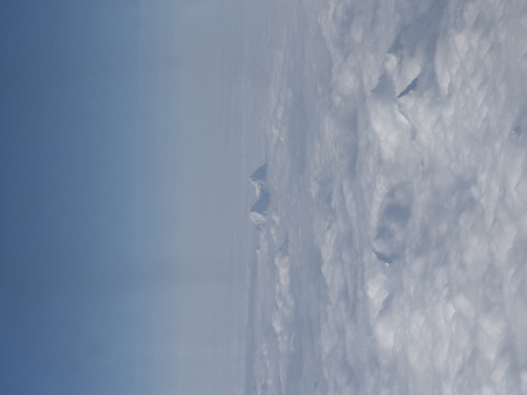 The image shows an aerial view of a landscape dominated by clouds. A clear, blue sky is visible on the left side of the image, transitioning into a thick layer of white and grey clouds on the right. Two snow-covered mountain peaks can be seen emerging from the clouds in the center of the image. Given the location of Gyazhugling town in China, it is likely the mountain range is the Himalayas. No people or other identifiable objects are visible.