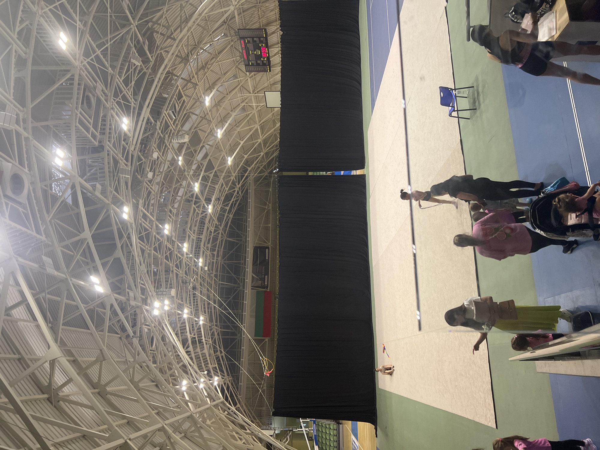 The photograph captures an indoor scene within a large sports hall, featuring a high, arched ceiling with an intricate exposed metal truss structure and numerous bright spotlights. The floor consists of distinct sections of light beige, green, and blue, forming courts.
Several individuals, predominantly women and children, are present in the hall. Some appear to be actively participating in or observing rhythmic gymnastics, with one person holding a skipping rope and another sitting on the floor surrounded by colorful clubs or ribbons. Other people are standing or seated, including one in a stroller, suggesting a mix of participants, coaches, and spectators. A lone blue plastic chair is placed on the beige court area.
Notable details include a large scoreboard high on a wall, displaying numbers such as "15.00" and "19.50," indicating a competitive event or training session. Large black curtains divide parts of the hall, possibly separating court areas. A Bulgarian flag, characterized by horizontal stripes of white, green, and red, is prominently displayed on a wall, confirming the location context of Plovdiv, Bulgaria. Above the flag, a dark banner with faint white text, possibly including "NEW BUILDING," is visible. The scene appears to be well-lit by artificial lighting, suggesting it could be daytime or evening.