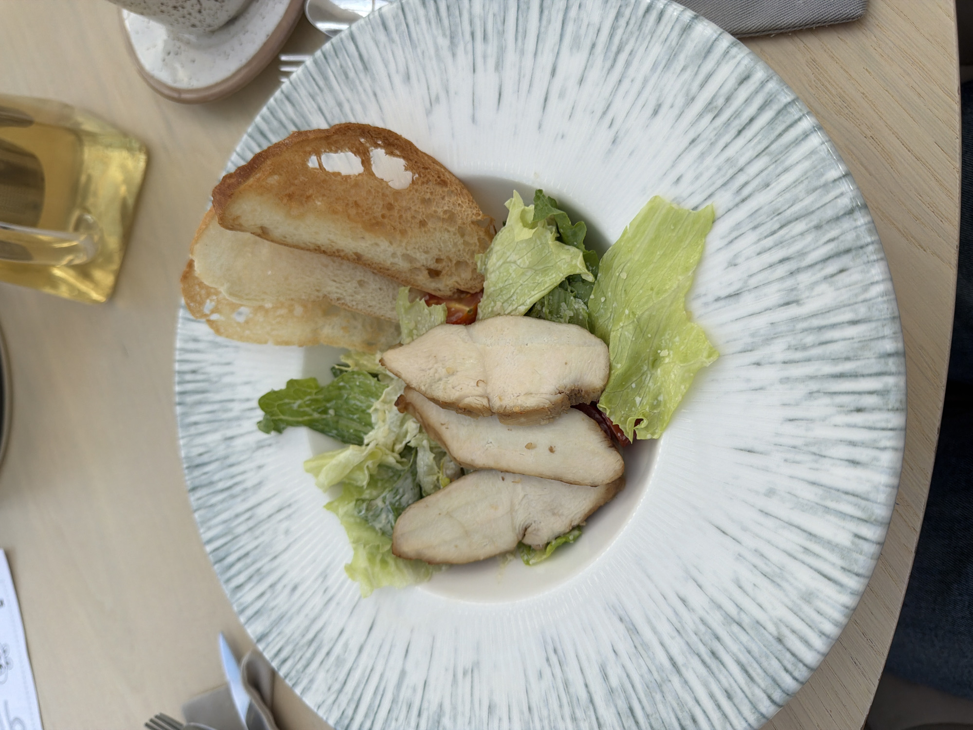 An overhead shot shows a meal presented on a distinctive white plate with radial grey stripe patterns. The main components of the dish include three slices of cooked chicken breast, fresh green leaf lettuce, and two pieces of toasted bread or large croutons. Small red pieces, likely diced tomatoes, are mixed with the lettuce, which appears to be lightly dressed.
The plate is set on a light-colored wooden table, suggesting an indoor dining environment such as a restaurant or cafe in Izhevsk, Russia. In the upper left corner, a portion of a textured ceramic cup or bowl is visible, along with a golden object that may be part of a drinking glass, reflecting light. To the bottom right of the plate, a grey napkin and the tines of a fork are partially visible. A white paper with a partial black character, resembling "b" or "6", is situated in the bottom left corner. The image focuses solely on the food and table setting, with no people visible.