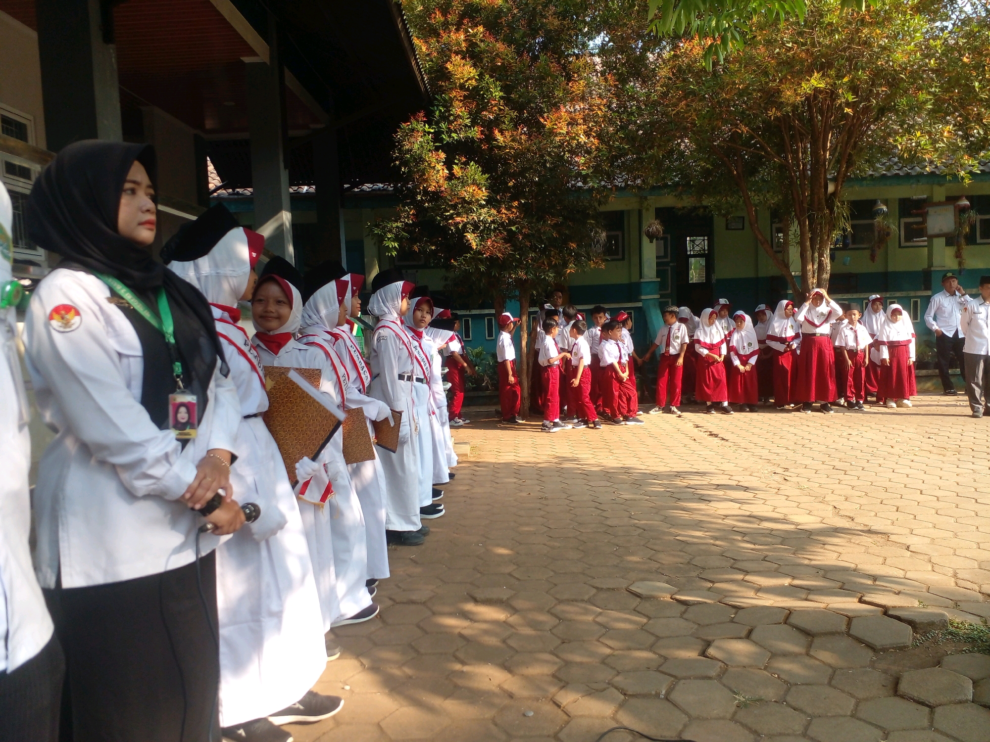 In Banyuputih, Indonesia, under a bright, sunny sky, a group of Indonesian students stand in formation on a paved courtyard outside a building. Many of the students are wearing white uniforms with red accents, and some are wearing red headscarves or caps. The students appear to be elementary or middle school-aged.
In the foreground, a woman in a black hijab and white uniform stands to the left, holding a microphone. She has a lanyard with an ID card around her neck. Behind her, a line of students in white uniforms with red trim are positioned. The students are mostly facing forward, with some looking towards the woman.
The background reveals a school building with a dark roof and a light-colored facade. Lush green trees with reddish-brown leaves are visible, casting dappled shadows on the ground. The courtyard pavement is made of hexagonal concrete blocks. Two men in white shirts are visible on the right side of the image, one of whom appears to be speaking on a cell phone. The overall scene suggests an outdoor school event or assembly.