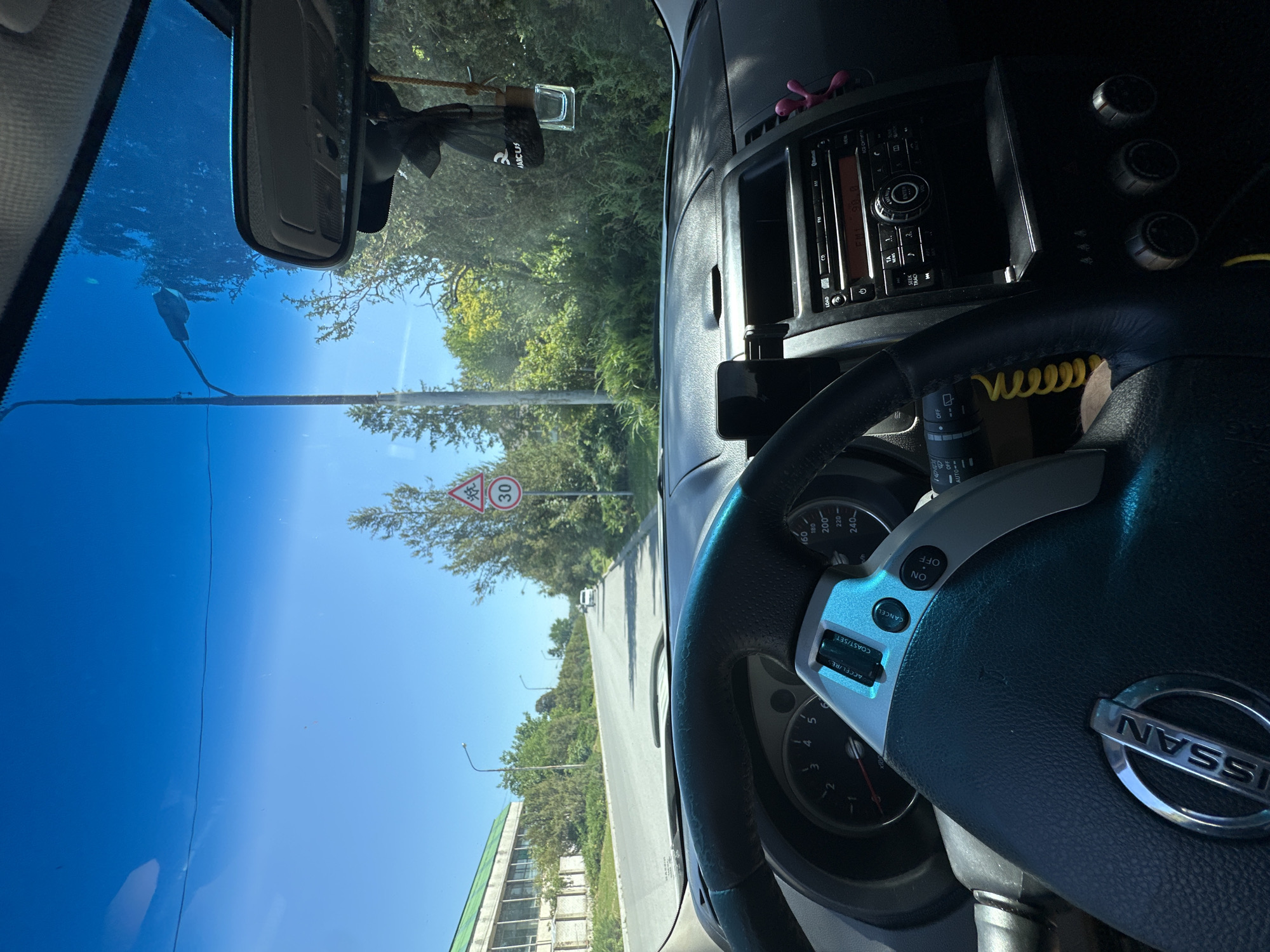 The image, taken from inside a Nissan car, shows a view through the windshield on a sunny day. The road ahead is paved and curves to the left. On the right side of the road, there is a triangular road sign with an image of children walking and the number "30" below it, indicating a speed limit of 30 kilometers per hour. Trees and foliage line the road. In the distance, a white car is visible. On the left side of the road, there is a large, low-rise building with a green roof. The interior of the car shows the steering wheel with a Nissan logo, the dashboard with a speedometer and other gauges, and the center console with a car stereo. A smartphone is mounted on the dashboard. A car air freshener hangs from the rearview mirror. The scene depicts a drive through an urban or suburban area, possibly Makiv, Ukraine, given the context.