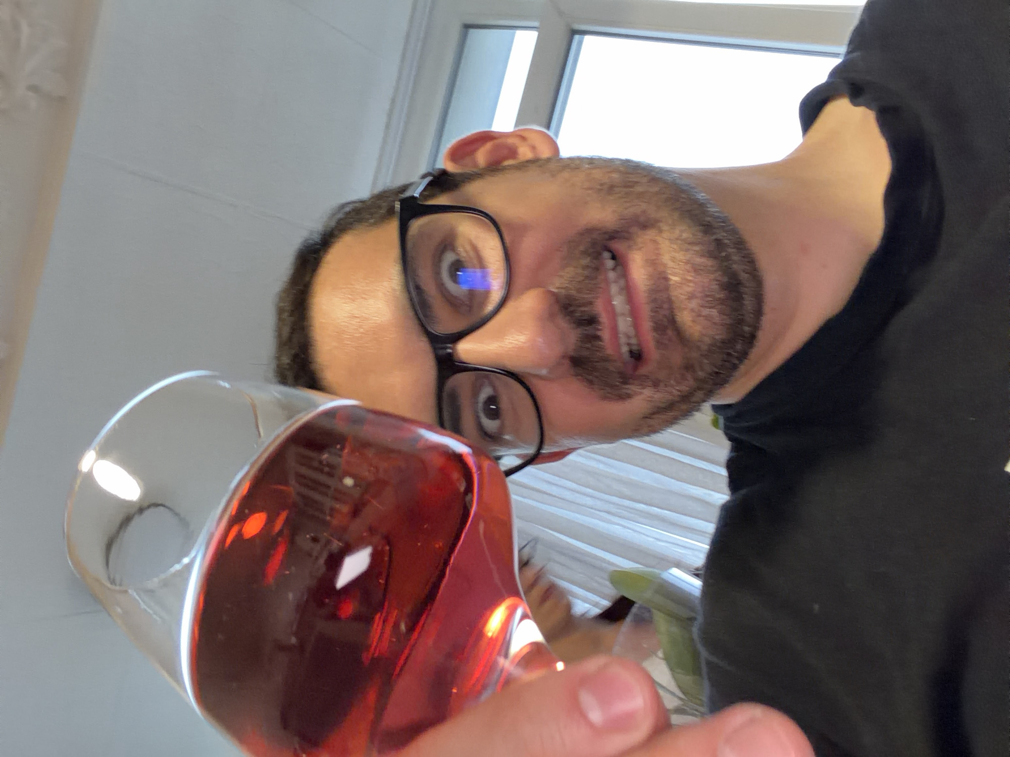 An eye-level, tilted selfie captures a man holding a glass of red liquid in the foreground. The man, seen from the chest up, has a dark beard and mustache, and is wearing black-framed glasses that reflect a blue light in his left eye. He looks directly at the camera with an open mouth, revealing his teeth, and an expression that appears surprised or slightly playful. His hair is dark and receding at the temples, and his skin tone is medium. He is wearing a black t-shirt or sweatshirt.
The glass he holds is thick-rimmed, clear, and filled with a translucent, dark red beverage, possibly wine or juice, which reflects light from the bright background. The background features a light-colored wall or ceiling on the left, and a window with white horizontal blinds or curtains on the right, through which bright daylight streams. The overall lighting suggests it is daytime. There is no visible text in the image. While the location context is Sofia, Bulgaria, no specific visual cues within the image confirm this location.