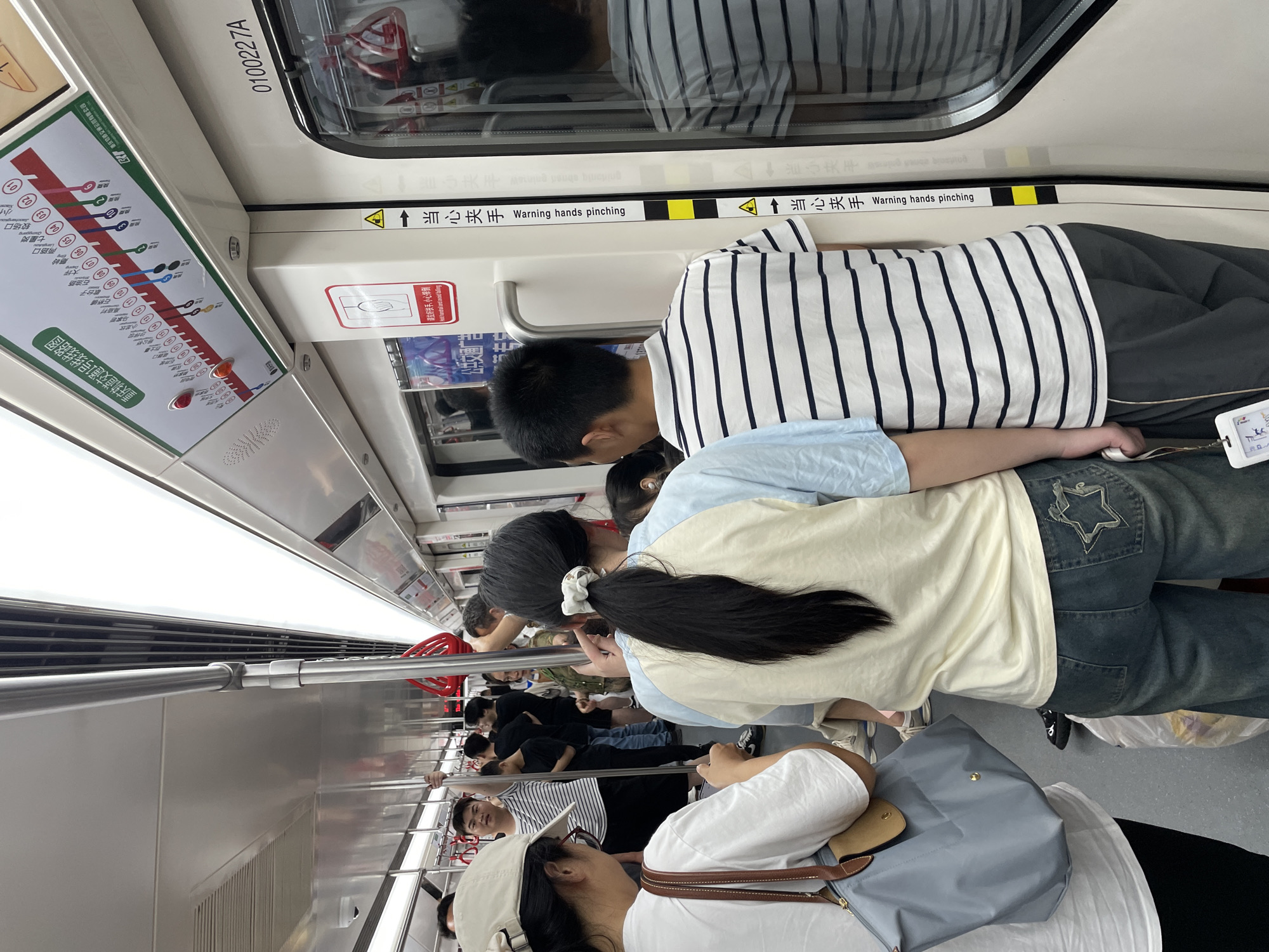 The image shows the interior of a crowded subway car. A route map for Chongqing Rail Transit Line 1 is visible, suggesting the location is Chongqing, China. Passengers are standing, some holding onto rails. Signage with Chinese characters and English reading "Warning hands pinching" is present. A person in a striped shirt and another with a ponytail are prominent in the foreground. A woman wearing a white shirt and hat is resting her head on a bag. The environment is brightly lit and modern.