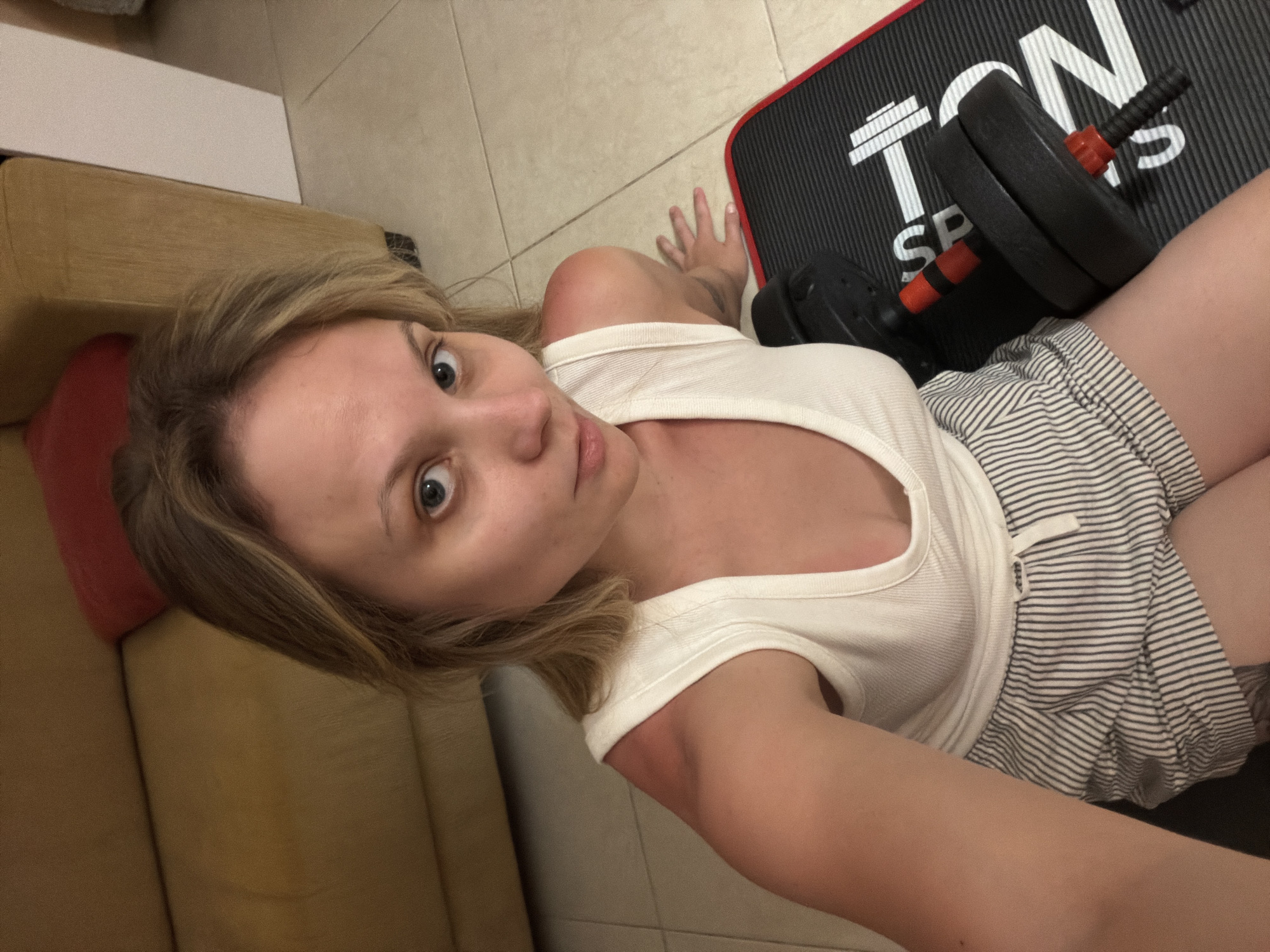 A young woman is captured in a close-up, high-angle selfie, lying on the floor in an indoor setting. She has fair skin, light brown hair, and bright blue-green eyes, looking directly into the camera with a neutral yet engaging expression. Her lips are slightly parted.
She wears a white, ribbed tank top and light-colored shorts with thin dark horizontal stripes. Her left arm extends towards the camera, suggesting she is holding the device for the selfie. Her right hand rests on the tiled floor. There is some redness visible on her shoulder and upper arm, which could indicate recent physical exertion or contact with a surface.
To her right, a black exercise mat is visible with white text that reads "TON SPORTS" and specifically "SP" from "SPORTS". A dumbbell with black weights and red handles/collars rests on the mat. The floor is covered in light beige rectangular tiles. To her left, a portion of a brown or tan sofa or armchair is visible, along with a red cushion. The indoor lighting suggests it is not primarily natural light. The scene depicts a casual moment, possibly related to exercise or relaxation, within a domestic environment in Tel-Aviv, Israel.