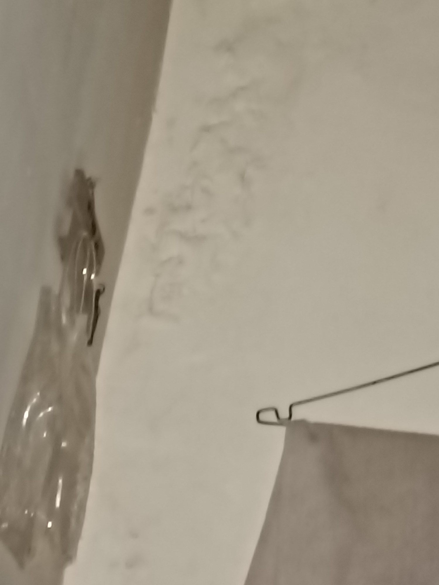 The image shows a close-up of a textured white wall, possibly a ceiling, with some discoloration and staining that suggests water damage or mold. To the left, a clear plastic bottle is partially visible, hanging by what appears to be a wire or string attached to a hook. The bottle is elongated and appears to contain a liquid or has been shaped to hold something. Further to the right, a thin, dark wire forms a hook shape, suspending a piece of light-colored fabric, possibly a towel or sheet. The fabric is draped over the wire and hangs down, filling the lower right portion of the frame. The overall impression is one of an indoor space, likely a bathroom or laundry area, given the presence of the fabric and the possible water damage on the wall. The lighting is dim, casting a yellowish hue over the scene. The image does not provide any specific details to identify its location within Kediri City, Indonesia.