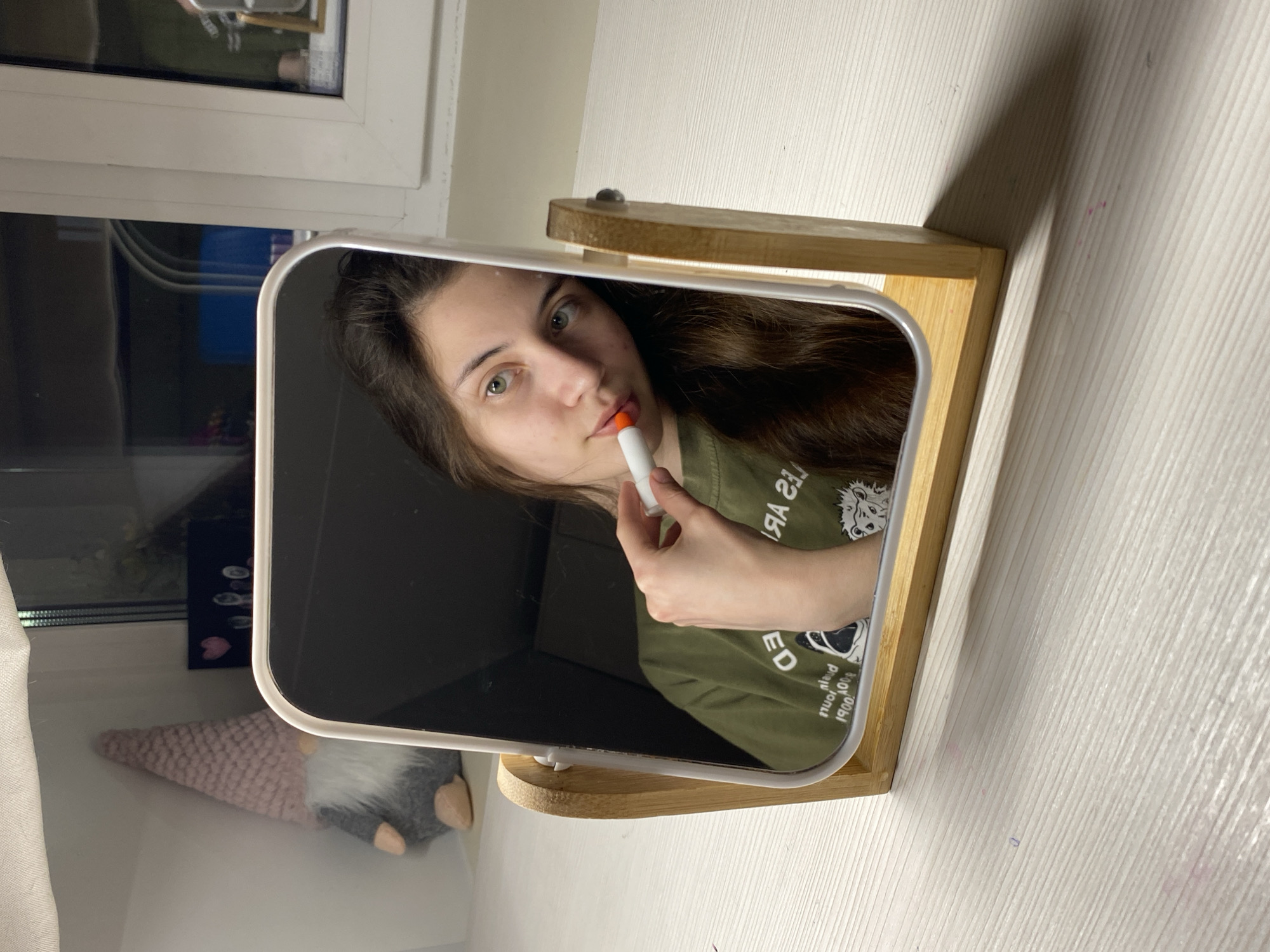 The image is an indoor shot, likely taken in a residential setting. A young woman is reflected in a small, square vanity mirror with a rounded top, set on a wooden stand. The mirror sits on a white surface with subtle horizontal graining, possibly a tabletop or shelf.
In the reflection, the woman is applying lipstick from a white tube with an orange top. She is wearing a green t-shirt with a design that includes a white animal graphic and text. Her hair is long, brown, and wavy. Her face is visible, showing fair skin, light eyes, and subtle makeup.
Outside the mirror's reflection, there is a window with a white frame in the background. On the windowsill, there is a small, decorative gnome-like figure with a pink knitted cap and a gray beard. There is also a small dark object decorated with pink hearts.
The setting appears to be a tidy, well-lit room with a simple, domestic atmosphere. The location is not discernible as Votkinsk, Russia, from the available visual information.