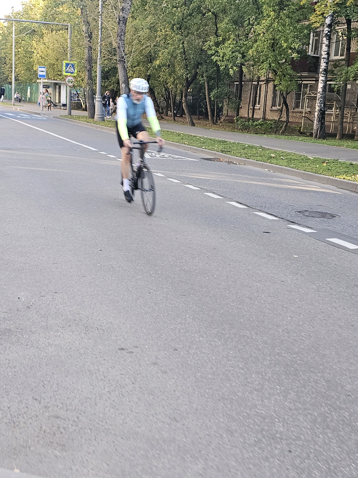 A man with a white helmet and grey beard cycles on a paved road in Khimki, Russia. He wears a blue cycling vest with bright yellow sleeves and black shorts. The cyclist is in motion, with his arms bent and his gaze directed forward. He is on the right side of the road, on a paved surface with white lane markings. To the left of the road, there is a bus stop with a blue sign and a triangular yellow sign depicting a pedestrian. Further to the left are trees and a brick building. The environment suggests an urban setting, possibly during the daytime with natural light. The image has a slight motion blur, indicating the movement of the cyclist or the camera.