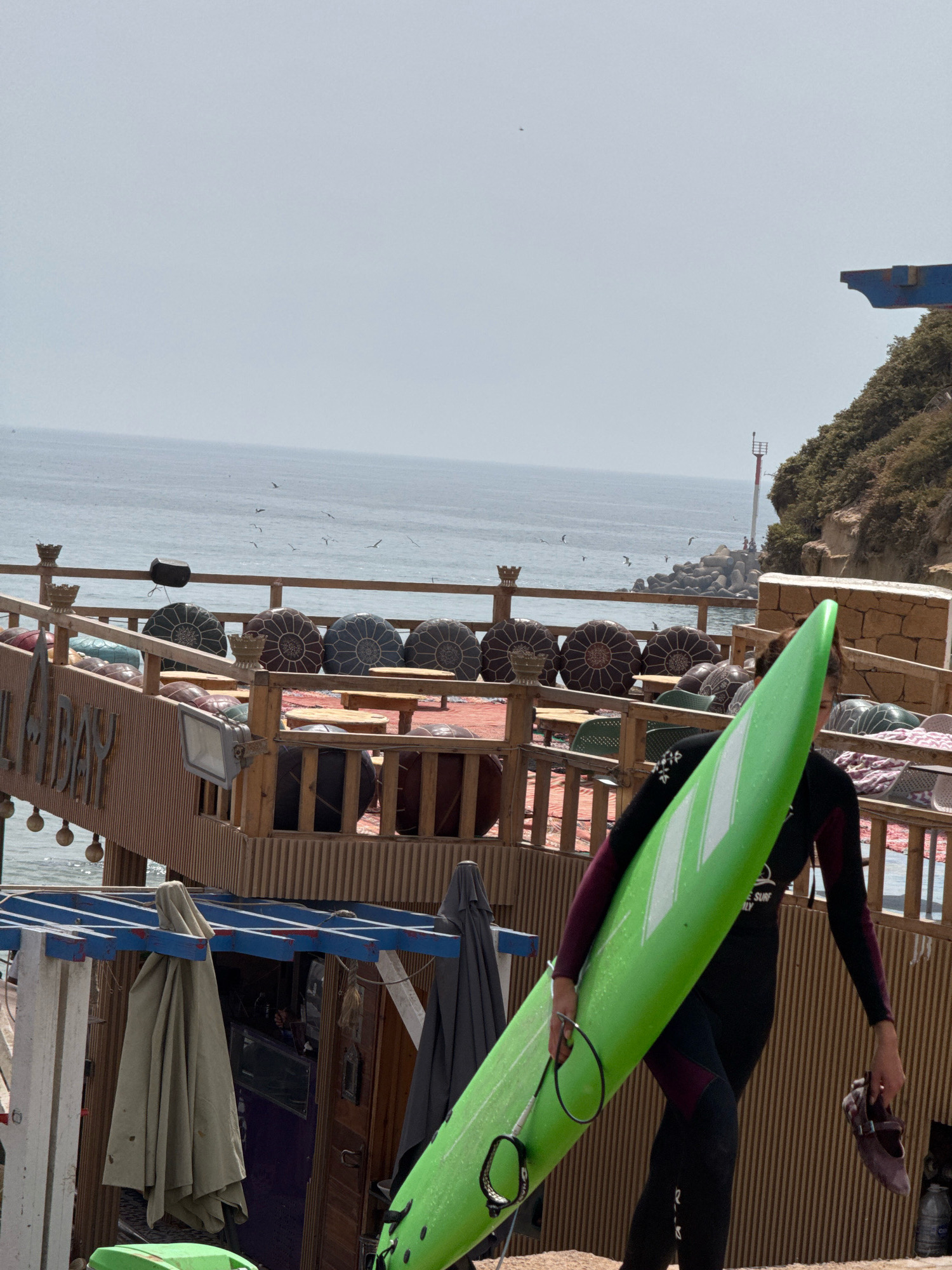 A person in a wetsuit walks on a beach in Imsouane, Morocco, carrying a bright green surfboard under their right arm. They are also holding a pair of brown flat shoes in their left hand. The background features a rustic wooden structure with tables and cushions, suggesting an outdoor seating area. Beyond that, the ocean stretches to the horizon, with a rocky coastline and a lighthouse visible on the right. Several seagulls are flying in the distance. The sky is overcast.