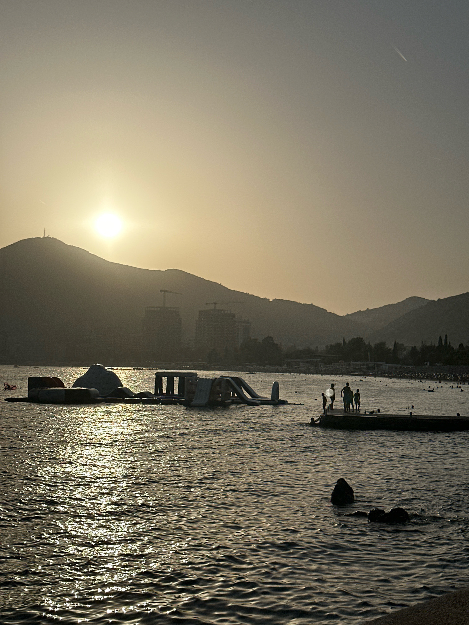 A wide shot captures a dusky, hazy sunset over the Adriatic Sea in Budva, Montenegro. The sun, a bright orb, is positioned low on the horizon, partially obscured by the silhouette of a large, tree-covered mountain that dominates the left side of the frame. The mountain has a telecommunications tower near its peak. Two unfinished high-rise buildings, under construction with cranes, are visible in the mid-ground against the mountain's slopes.
In the foreground, the sea is choppy, with sunlight glinting off the surface, creating a shimmering, textured effect. Several inflatable water toys, including a large, dome-shaped structure and slides, are anchored in the water. A wooden pier juts out into the sea on the right side, where a group of silhouetted figures, appearing to be children and adults, stand. One person is in the water near the pier. In the distance, the shoreline is dotted with many more silhouetted people, suggesting a crowded beach. The overall atmosphere is that of a warm, late summer evening at a popular seaside resort.