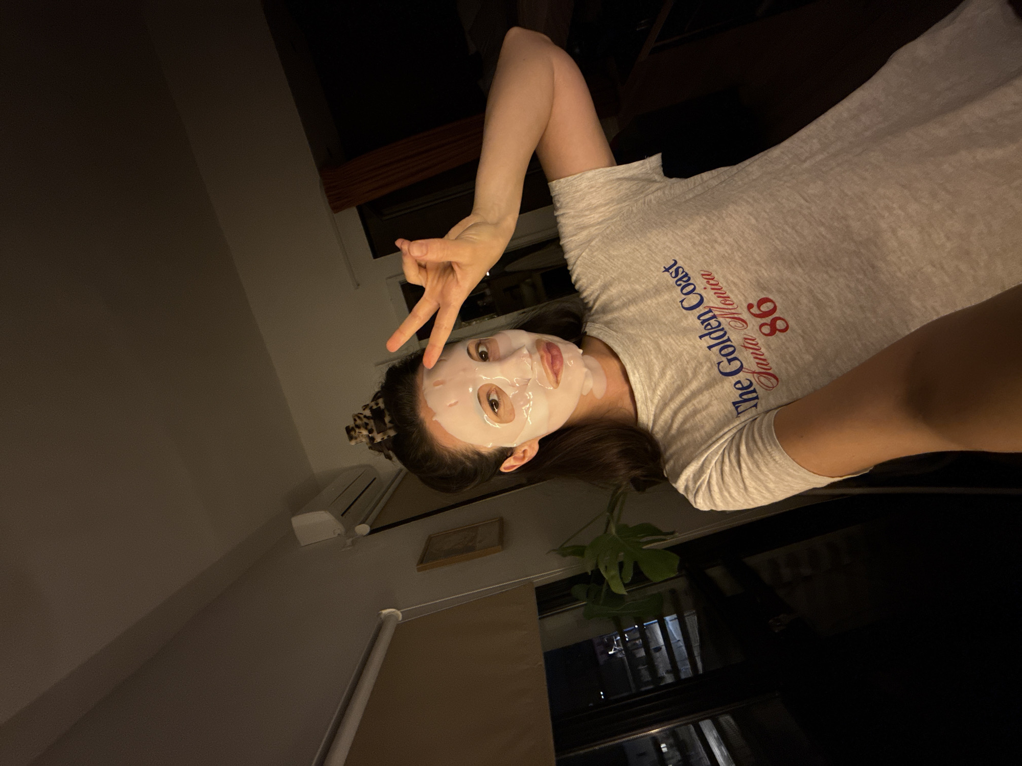 A woman is posing for a selfie indoors. She has a white face mask on and is holding up a peace sign with her left hand. She is wearing a gray t-shirt with the words "The Golden Coast, Santa Monica '86" printed on it. Her hair is pulled back with a leopard-print hair clip.
The background is dimly lit and includes a window, a potted plant, an air conditioner, and a framed picture on the wall. The woman is inside an apartment or home, but there's no indication of it being in Tel-Aviv, Israel.