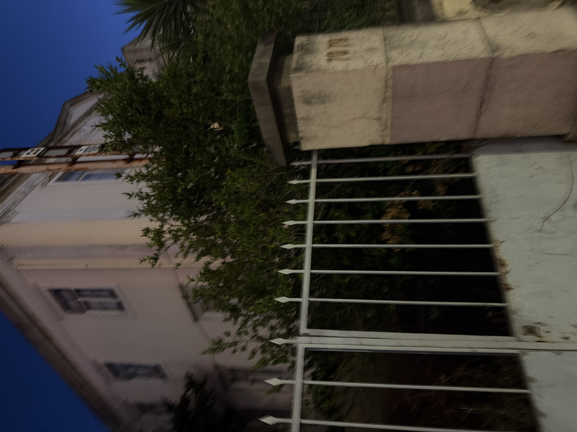 The image shows a two-story building with white stucco walls and dark-framed windows at dusk in Cascais, Portugal. A white metal fence with pointed finials runs across the foreground, partially obscuring a weathered white wall. To the right of the fence, a stone pillar is visible, featuring the numbers "70 79" etched into its surface. Lush green foliage borders the property, with a tall palm frond visible on the right edge of the frame. The sky is a deep, clear blue, indicating twilight. A rusty metal pipe runs vertically along the side of the building.