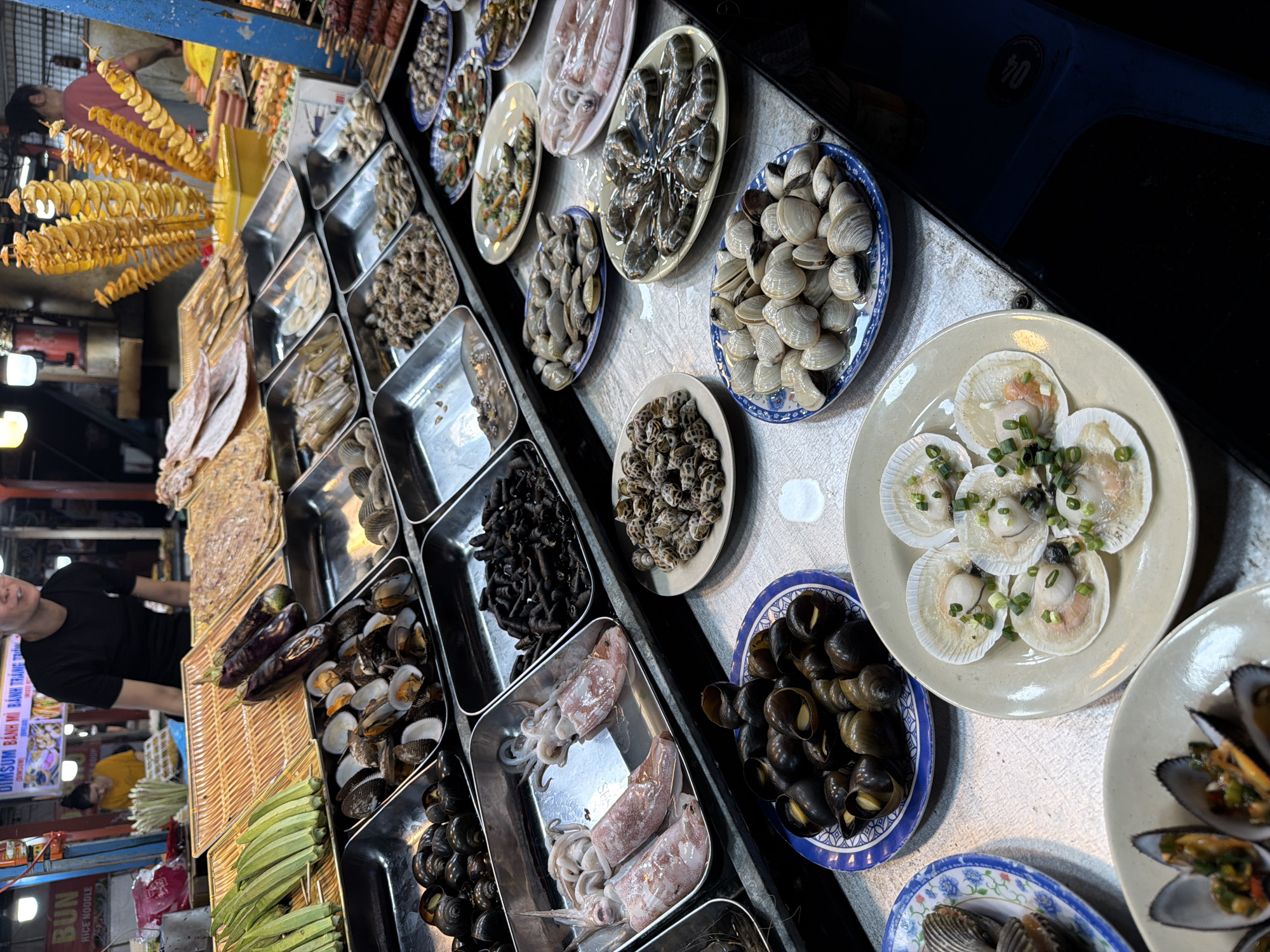 The image shows a street food stall in Đà Nẵng, Vietnam, displaying a variety of fresh seafood and vegetables. In the background, two vendors are visible behind a counter, one tending to food and another, a man in a black t-shirt, is looking towards the camera. The stall features several metal trays and woven baskets filled with food. Among the items are green okra on skewers, dark purple eggplants, dried seafood, various types of snails, clams, scallops, squid, and shrimp. Some dishes are presented on ceramic plates, such as scallops with green onions, clams, and small snails. A display of spiral-cut potatoes on skewers is also prominent. The stall is situated outdoors, with overhead lighting and other market stalls visible in the distance. The overall scene captures a bustling atmosphere typical of a Vietnamese night market or food street.
