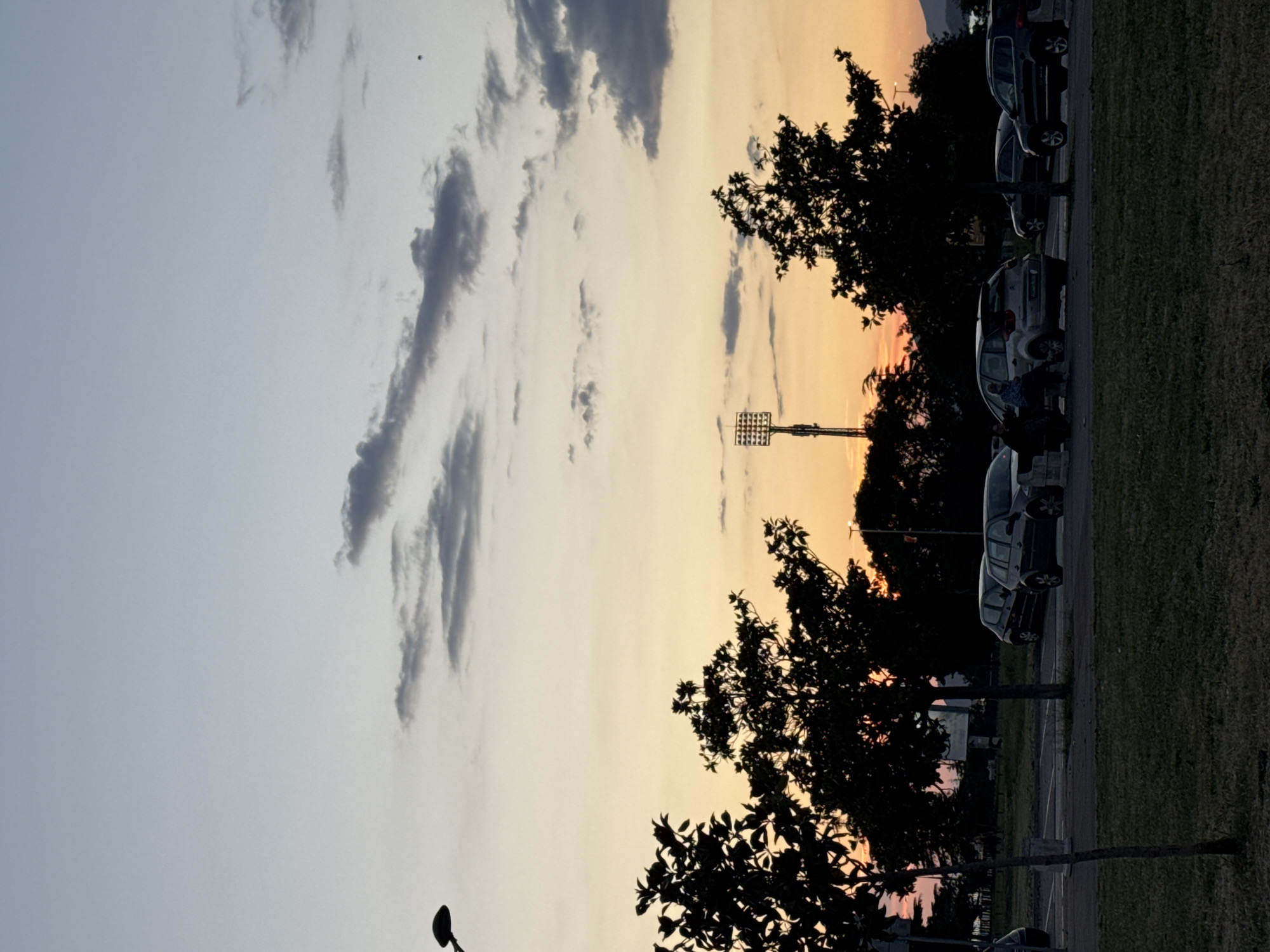 The image shows a parking lot with several cars parked alongside a grassy area, with trees lining the edge. The sky is a gradient of colors from light blue to orange, with clouds visible. A tall stadium light pole stands in the distance. The photo was taken in Bar, Montenegro.