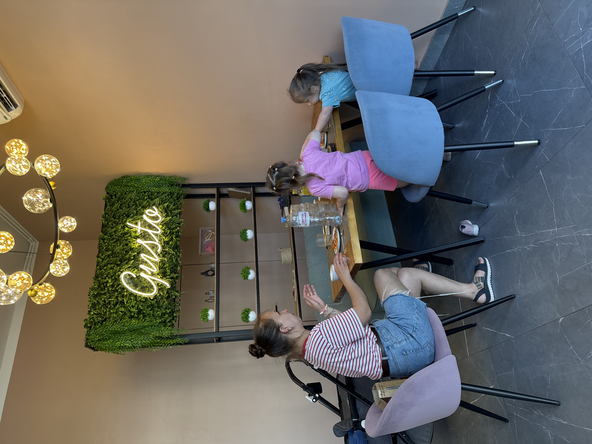 A woman sits at a wooden table in a restaurant with two young girls. The woman, wearing a striped t-shirt and denim shorts, is gesturing with her hands. One girl, in a pink t-shirt and shorts, is reaching across the table. The other girl, in a blue t-shirt, is also at the table. A bottle of water and a slice of pizza are visible on the table. The restaurant has a wall with artificial greenery and a neon sign that reads "Gusto." There are two plush chairs, one lavender and two blue, and a stroller in the background. The floor is dark grey with white marbling. The overall ambiance suggests a casual dining setting in a restaurant located in Patrusheva, Russia.