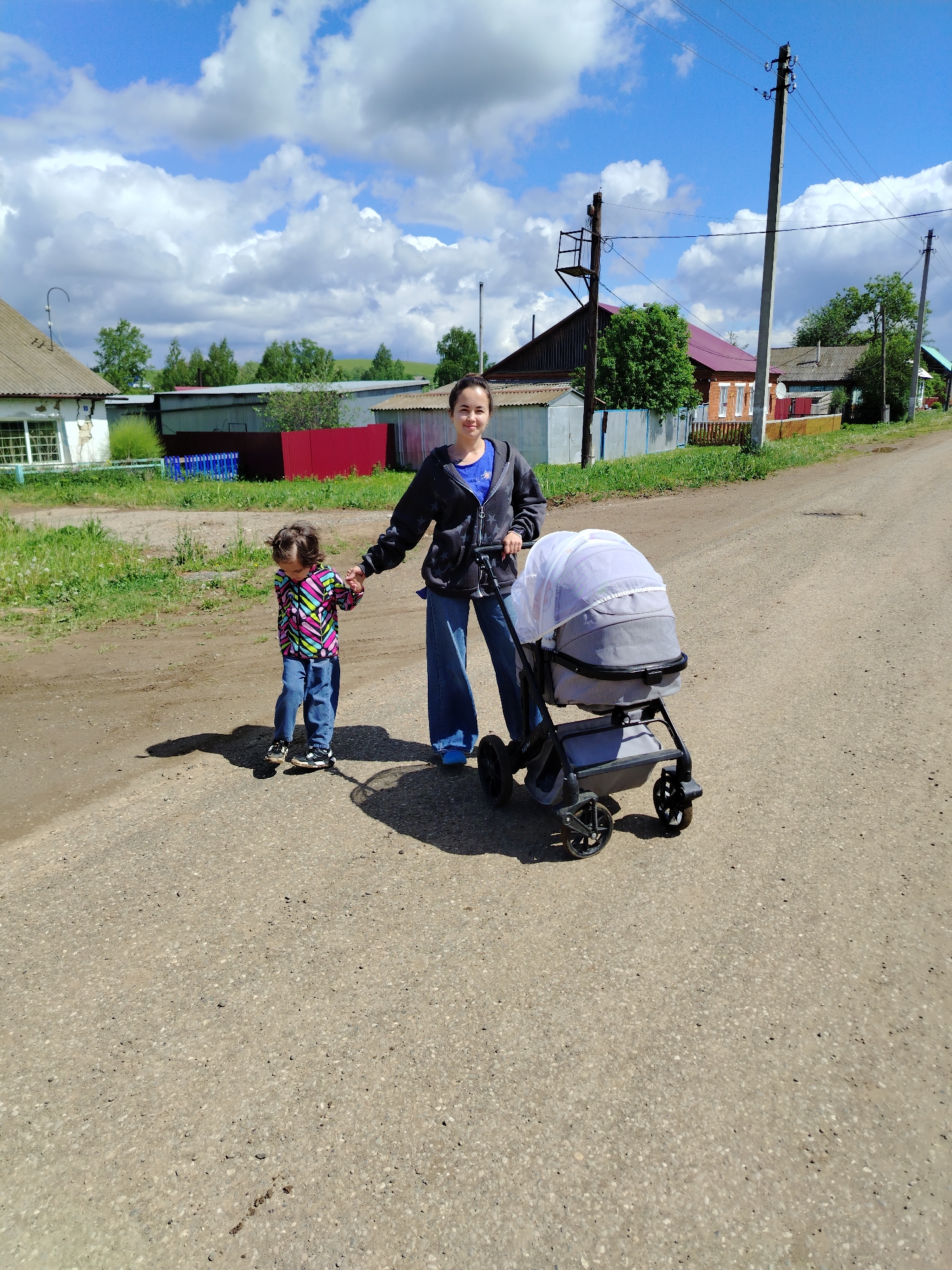 The image shows a woman, a child, and a stroller on a dirt road. The woman is fair-skinned with dark hair pulled back. She wears a blue t-shirt, a dark zip-up jacket with white stars, and wide-legged blue jeans. She is holding the hand of a young child. The child wears a multi-colored patterned jacket, blue jeans, and black-and-white sneakers. The child's hair is curly and dark. Next to the woman is a gray stroller with a white mesh cover. The dirt road stretches into the distance, with houses and foliage visible on either side. Utility poles are also visible. The sky is blue with scattered white clouds. Based on visual cues, this photo was taken in Гожан, Russia.