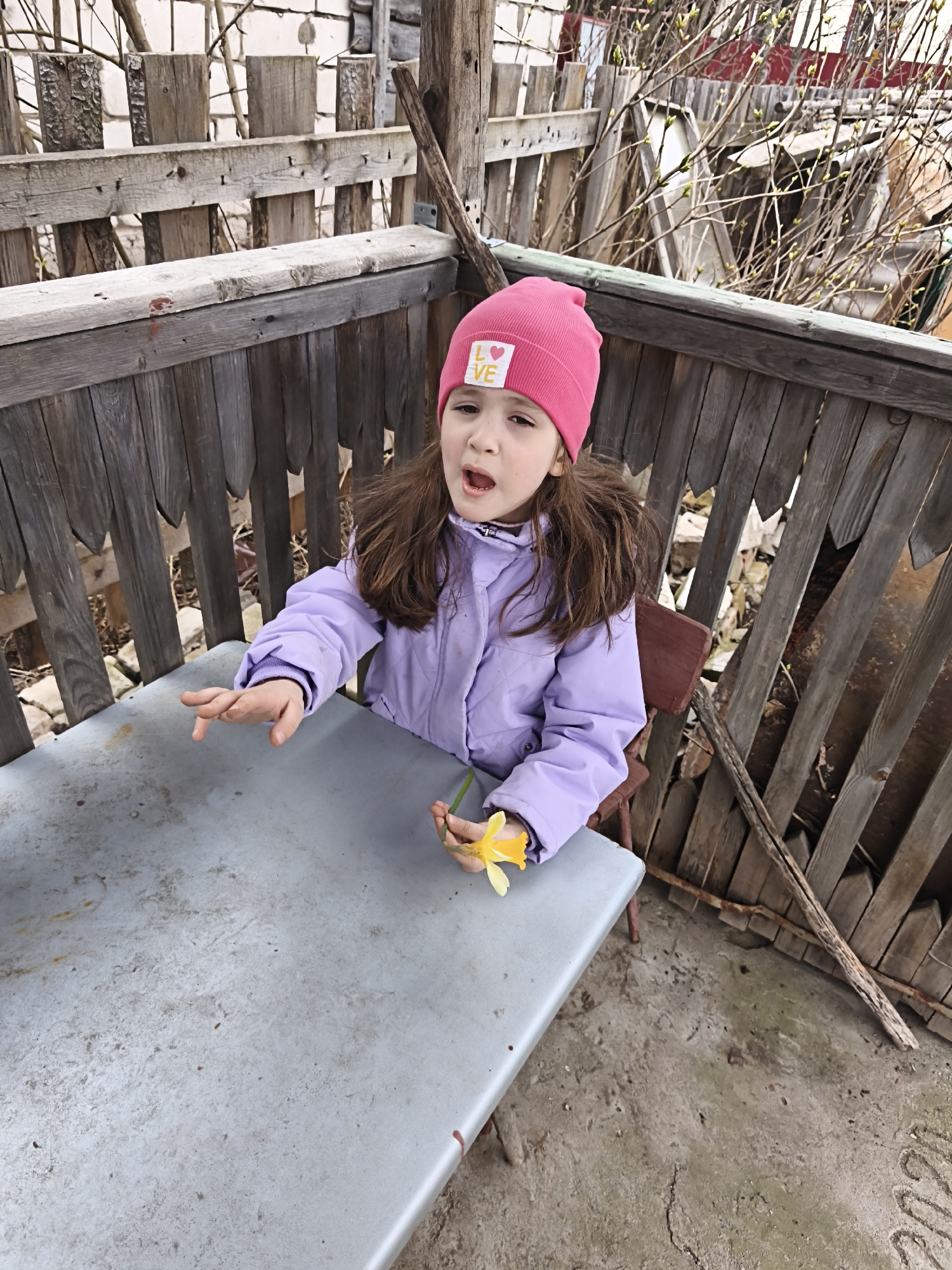 A young girl sits at a gray table on a dilapidated porch in Прикамский, Russia. She wears a pink hat with a "LOVE" patch and a lavender jacket. She is holding a yellow daffodil in one hand and gesturing with the other. The porch has a weathered wooden railing, and the ground is bare. In the background is a mix of wooden structures, including fences and possibly a shed, along with bare tree branches.