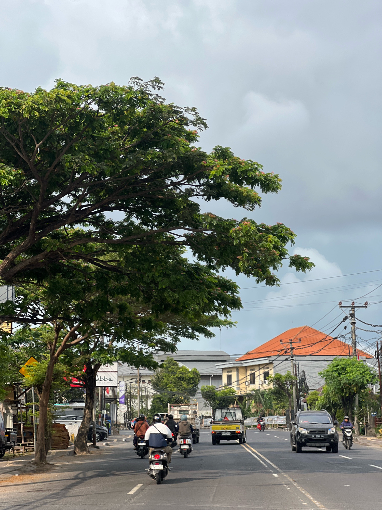 The image depicts a street scene in Denpasar, Indonesia, on a cloudy day. A large, leafy tree with some pink blossoms dominates the left side of the frame. The road is paved and marked with double white lines, indicating two lanes of traffic. Several motorbikes are visible, with riders wearing helmets and casual clothing, moving in the same direction. A yellow utility vehicle with "maxim" written on its side is also present. Further down the road, a black SUV is driving, and another motorbike can be seen in the distance. On the right side of the road, there are utility poles with electrical wires and a building with an orange tiled roof. In the background, other buildings and more trees are visible. The overall atmosphere suggests a typical urban environment with moderate traffic.
