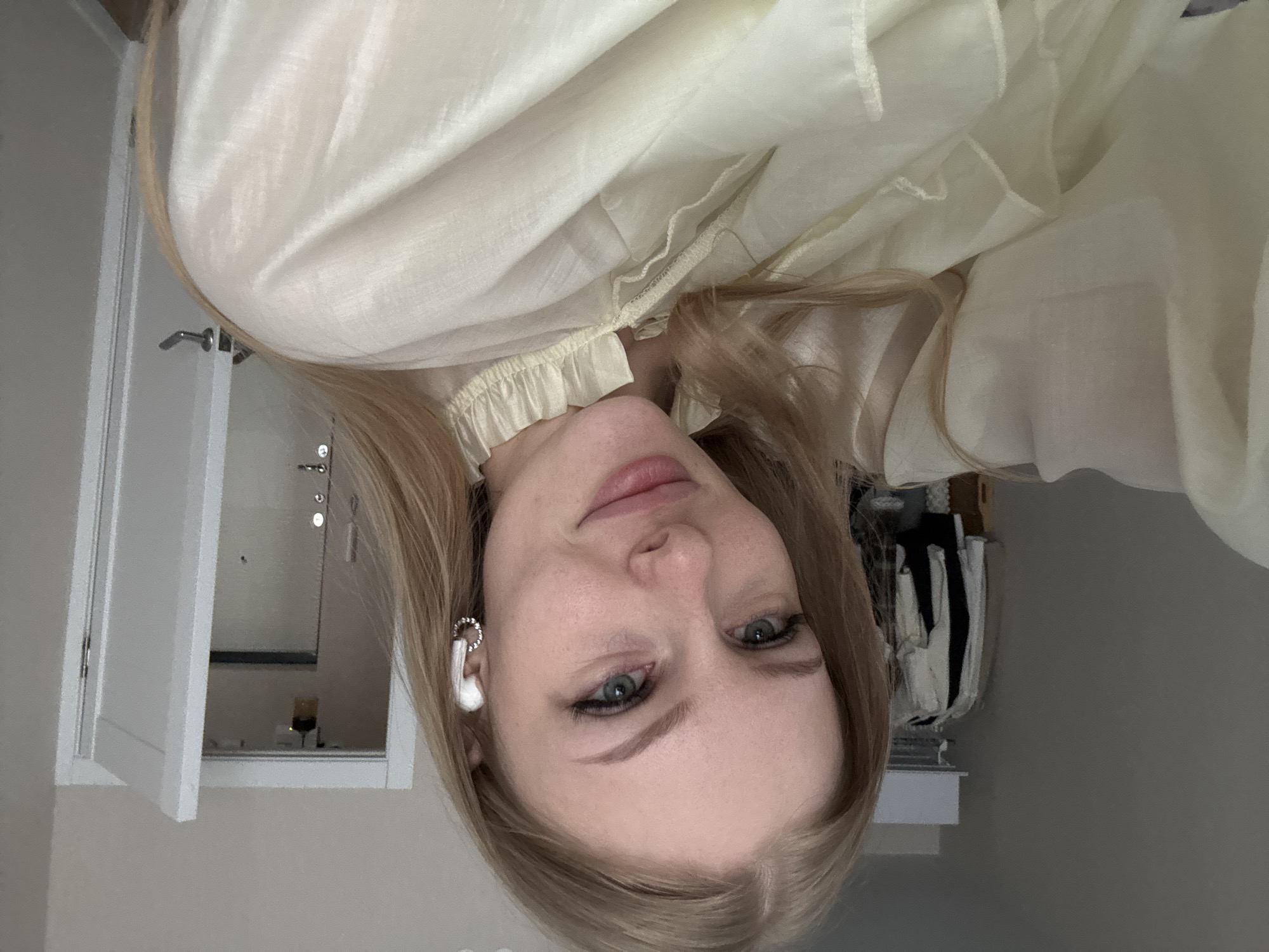 A young Caucasian woman with blonde hair and blue eyes is pictured in a selfie. She is wearing a light yellow ruffled blouse and white wireless earbuds. Her left arm is visible, and she is wearing a silver hoop earring on her left ear. The background appears to be a bedroom with a closet containing clothing and a partially open door leading to what looks like an apartment hallway. The location is stated to be Perm, Russia.