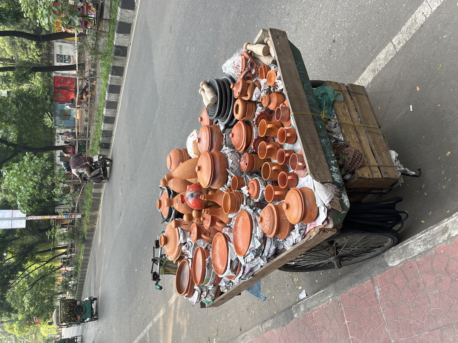 This medium-shot photograph, taken on a sunny day in Dhaka, Bangladesh, captures a bustling street scene centered around a vendor's cart laden with pottery. The image is oriented with the road running vertically through the middle.
In the foreground, a large, wooden handcart occupies the right side, packed with numerous terracotta items. These include various pots, bowls, cups, plates, and decorative pieces, predominantly in a natural reddish-brown earthenware color, some featuring carved patterns. The pottery is carefully arranged, often cushioned with newspaper or white fabric. On the rear of the cart, wooden planks and bundles of dark, coiled material (possibly hoses or ropes) are visible. A spoked wheel of the cart is at the bottom right. The sidewalk next to the cart is paved with reddish-brown tiles.
The road itself is asphalt, with a white line marking its edge and a black-and-white striped curb separating it from a narrow, grassy median. On the road, a person on a motorcycle, wearing a dark shirt and helmet, rides in the midground. Further down the road, a green auto-rickshaw is also visible. Beyond the road, on the left, a green area with numerous trees and foliage extends into the background. Behind these trees, a wall or fence features colorful murals or graffiti, some of which appear to have Bengali script, though it's illegible due to distance. A white sign with what looks like Bengali text and the number "500" is also faintly visible in the upper left background. The bright lighting and visible shadows suggest it is daytime, likely mid-morning or afternoon, with clear weather.