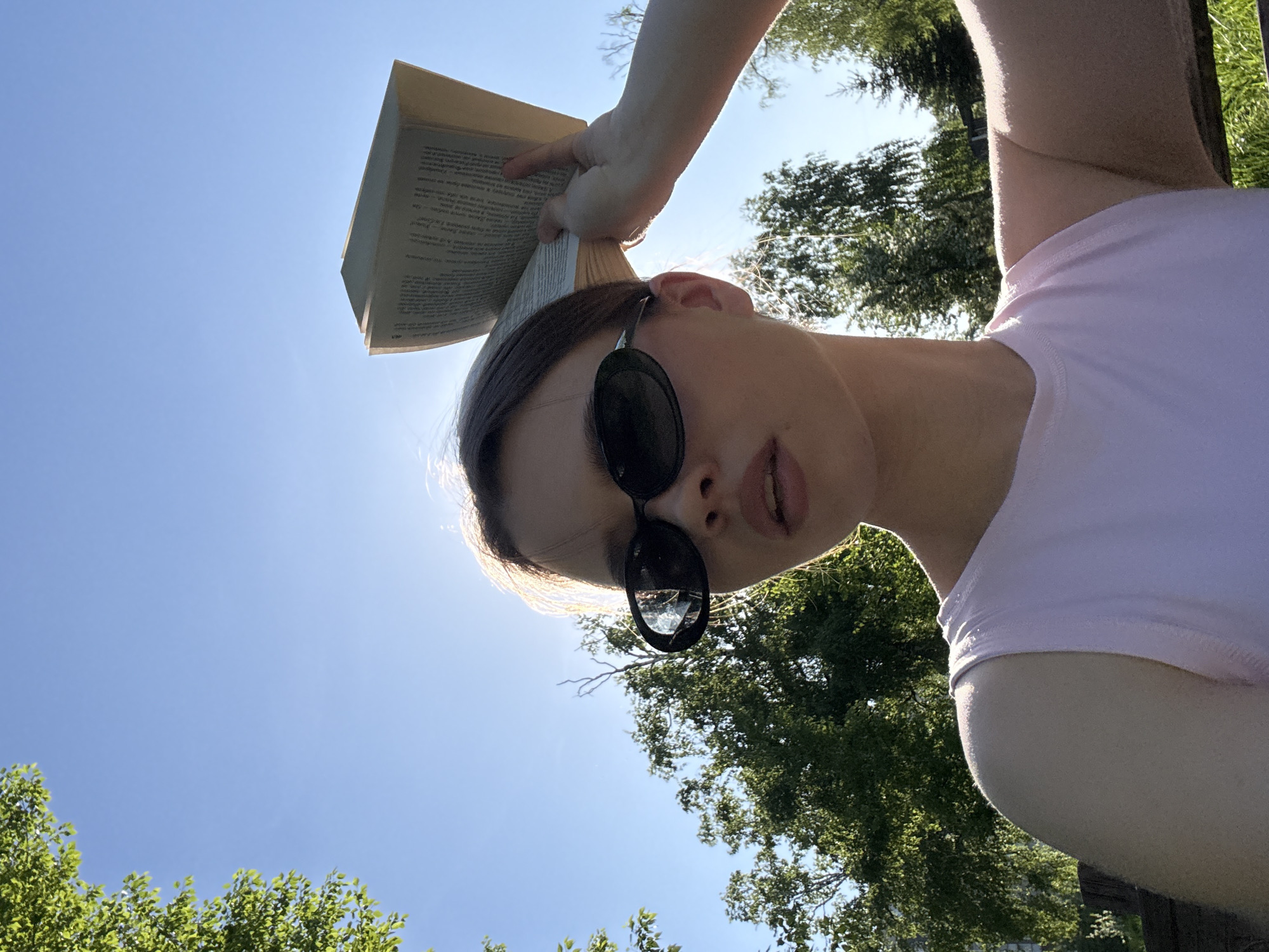 The image captures a person outdoors on a bright, sunny day in Krasnoyarsk, Russia, viewed from a low-angle perspective looking upwards. The subject is a person, likely female, with light skin and brown hair, wearing dark, round sunglasses and a light pink sleeveless top.
A book is held open directly above her head, possibly to shield her face from the sun. The visible text on the book's pages is in French, with discernable words such as "l'oeuvre," "conçues," and "le corps." A faint page number, possibly "46" or "97," can be seen. Her left hand is visible, holding the book open.
The time of day is clearly mid-day or early afternoon due to the intense sunlight and clear blue sky. The sun is positioned behind and slightly above the person, creating a bright halo effect around her hair and the book. Green tree foliage is visible in the background, framing the clear sky. The person's head is tilted back, and she is looking down towards the camera with her mouth slightly open. The scene conveys a relaxed, sunny outdoor setting.
