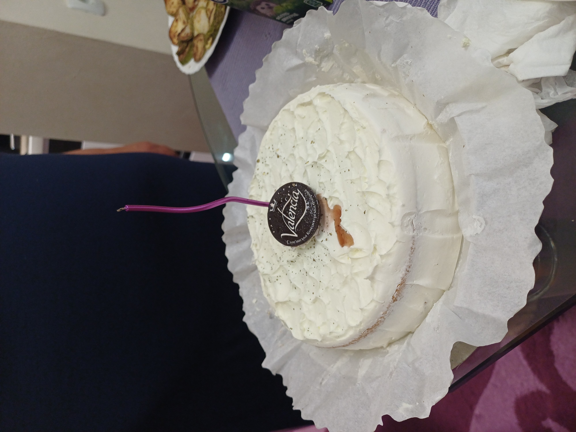 A birthday cake with white frosting, a single pink candle, and a dark circular chocolate decoration with the text "Valencia" is on a table. The cake is on a white paper liner. A plate of sliced zucchini is in the background to the right of the cake. The table is covered with a purple cloth. A person wearing a dark blue shirt is partially visible on the left side of the image. The setting appears to be a home environment in Odesa, Ukraine.