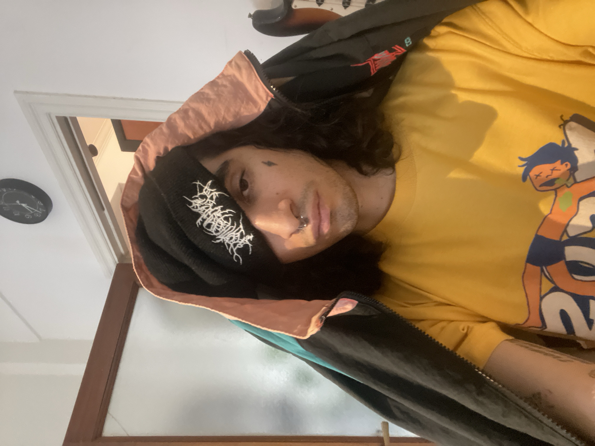 A person is seen from a slightly high, canted angle. They wear a yellow t-shirt with a cartoon graphic, a black beanie with white lettering, and a black jacket with a peach-colored hood lining. A nose ring is visible. There is a black tattoo of a small lightning bolt on their face, near their eye. Their hair is long, dark, and curly. A guitar hangs on the wall behind them. A doorway with a clock on the wall above is visible behind the person. A window is also visible behind the person. It is not possible to determine a specific location in Buenos Aires, Argentina from this image.