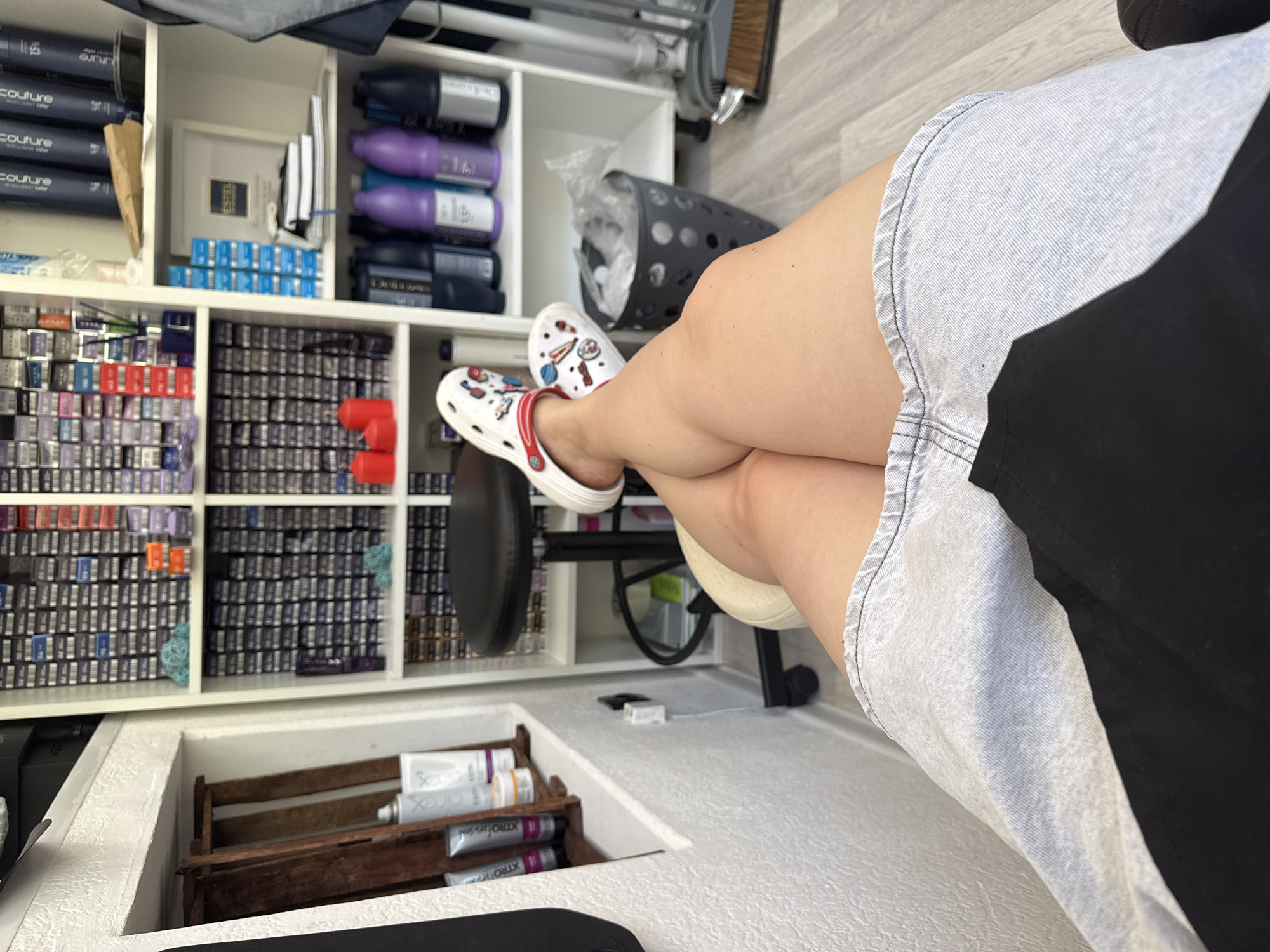 A person with crossed legs, wearing white crocs with charms, sitting in a white chair in what appears to be a salon in Chaikovsky, Russia. Shelves behind them contain hair products and supplies. There are tubes of product stored in a wooden crate below the shelves.