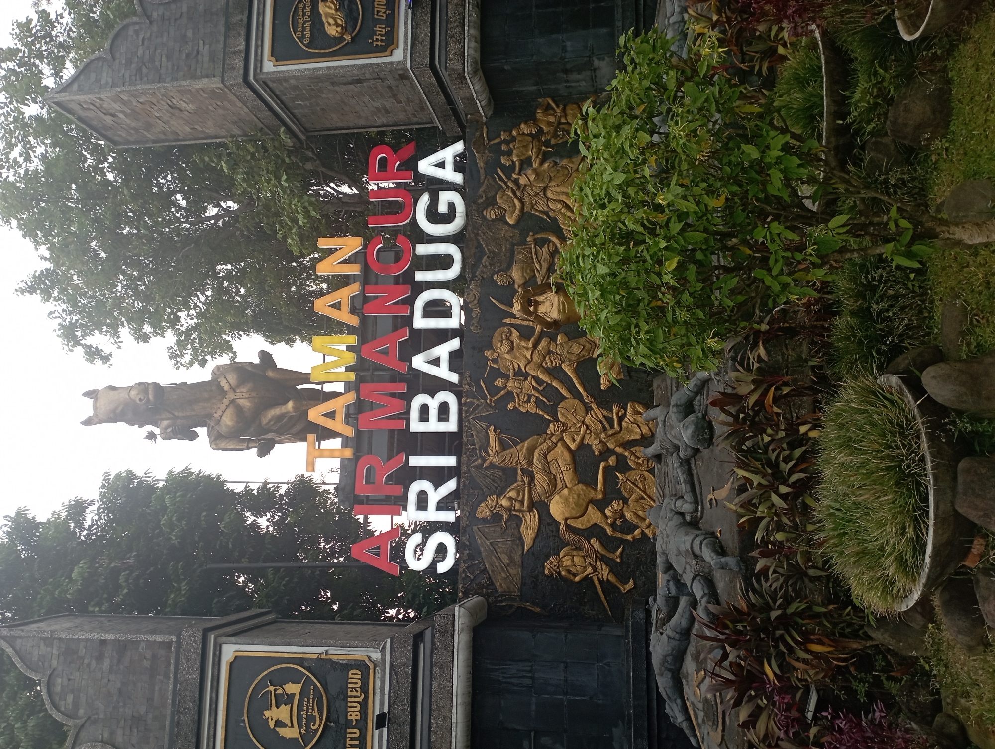 The image depicts the entrance to Taman Air Mancur Sri Baduga, a park located in Purwakarta, Indonesia. The central feature is a large monument with a statue of a warrior on horseback at the top, possibly representing a historical figure. Below the statue, the park's name is prominently displayed in large, colorful letters: "TAMAN AIR MANCUR SRI BADUGA". The monument is adorned with bas-relief sculptures depicting scenes of battle, featuring warriors on horseback and fallen figures. The surrounding area is landscaped with trees, bushes, and ornamental plants, and the overall scene is presented in an overcast or hazy lighting condition. There are no people visible in the image.